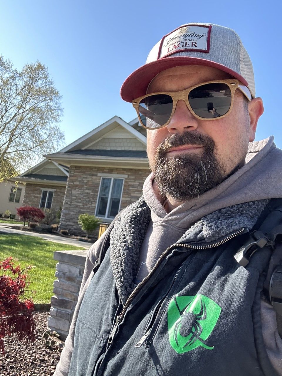 Man in sunglasses and hat, in front of a house, wearing a vest with a green logo.