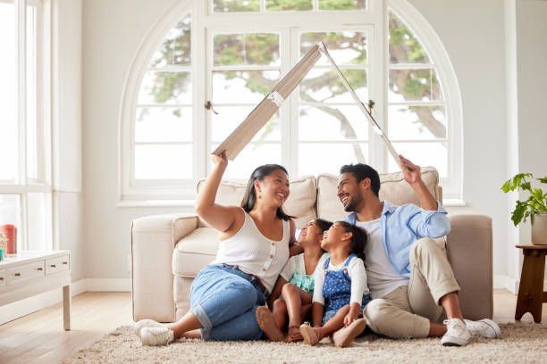 A family is sitting on the floor in a living room holding a cardboard house.