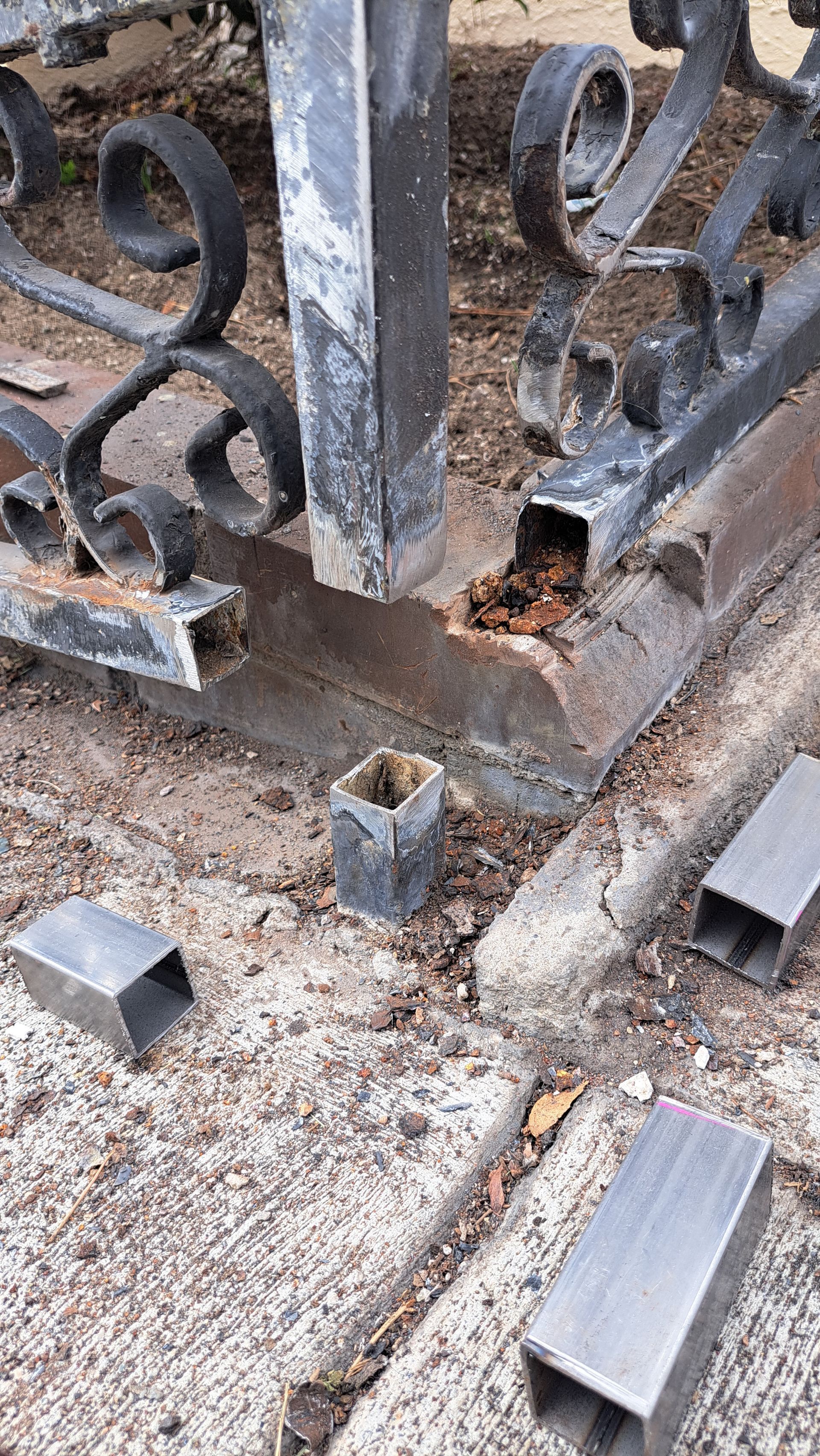 A broken metal gate with square posts and ornate scrollwork, resting on a concrete base with detached metal pieces scattered.