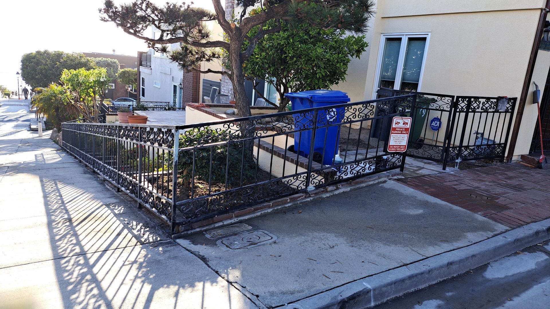 Black wrought-iron fence along a sidewalk in front of a house. A blue trash bin is visible behind the fence.