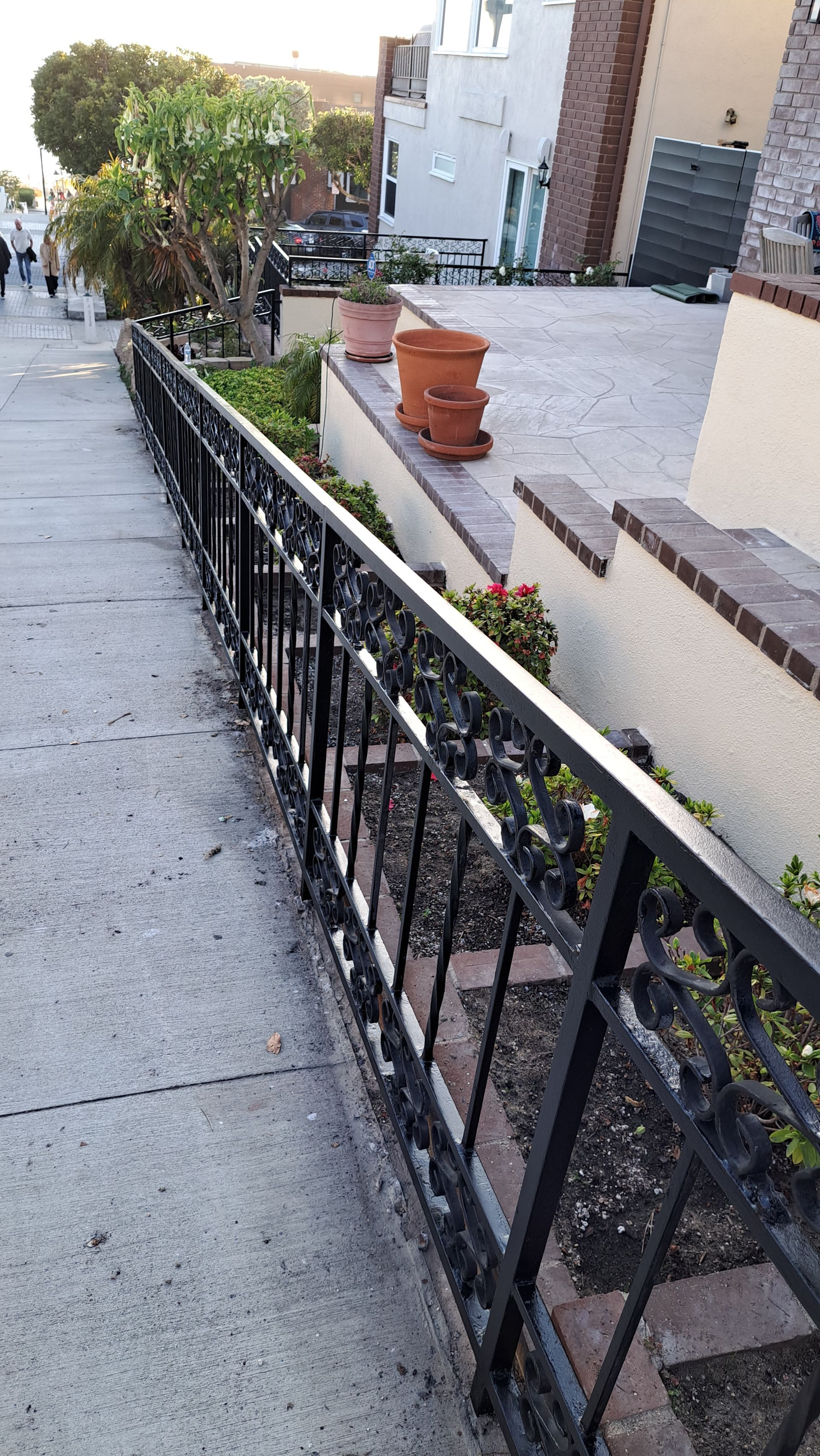 Black wrought iron fence along a sidewalk with plants and potted flowers.