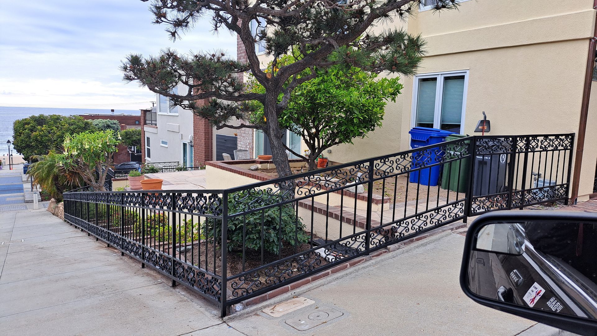 A house with a decorative wrought iron fence on a sloped sidewalk, a small tree, and a view of the ocean.