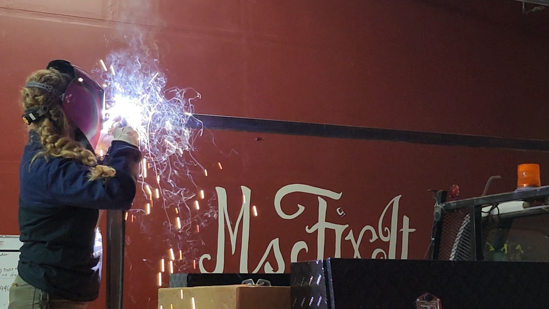 Person welding a metal bar; sparks fly. Red wall has white text reading 
