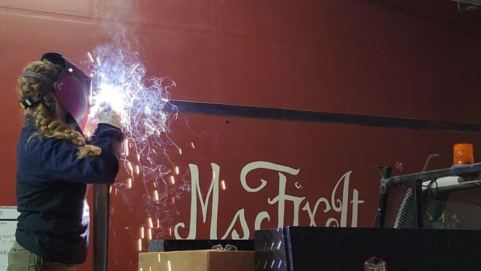 Person welding a metal bar; sparks fly. Red wall has white text reading 