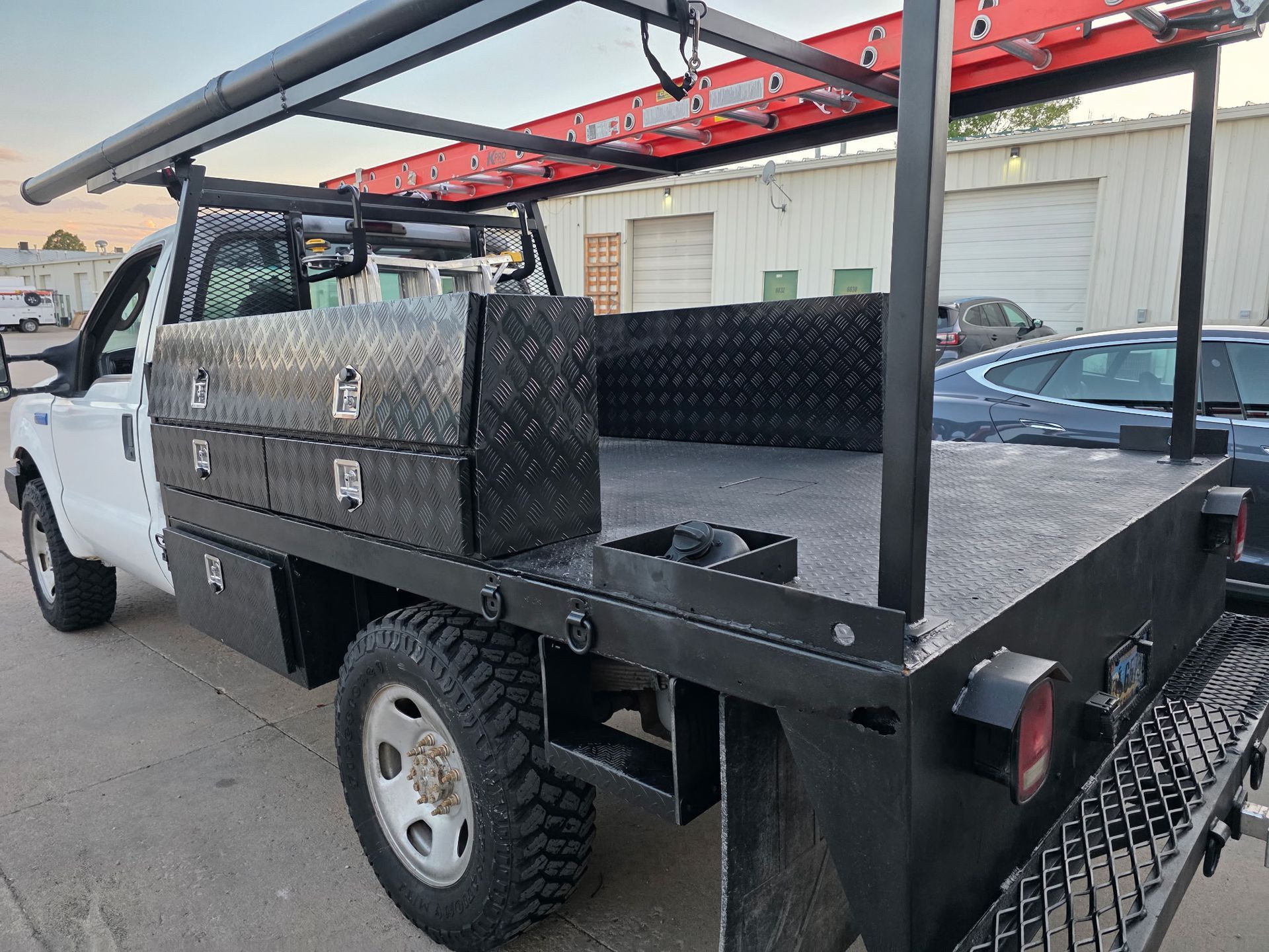 White work truck with a black utility bed and ladder rack, parked outside.