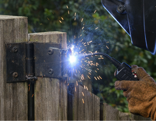 A person welds a hinge onto a wooden fence. Sparks and bright light are visible.