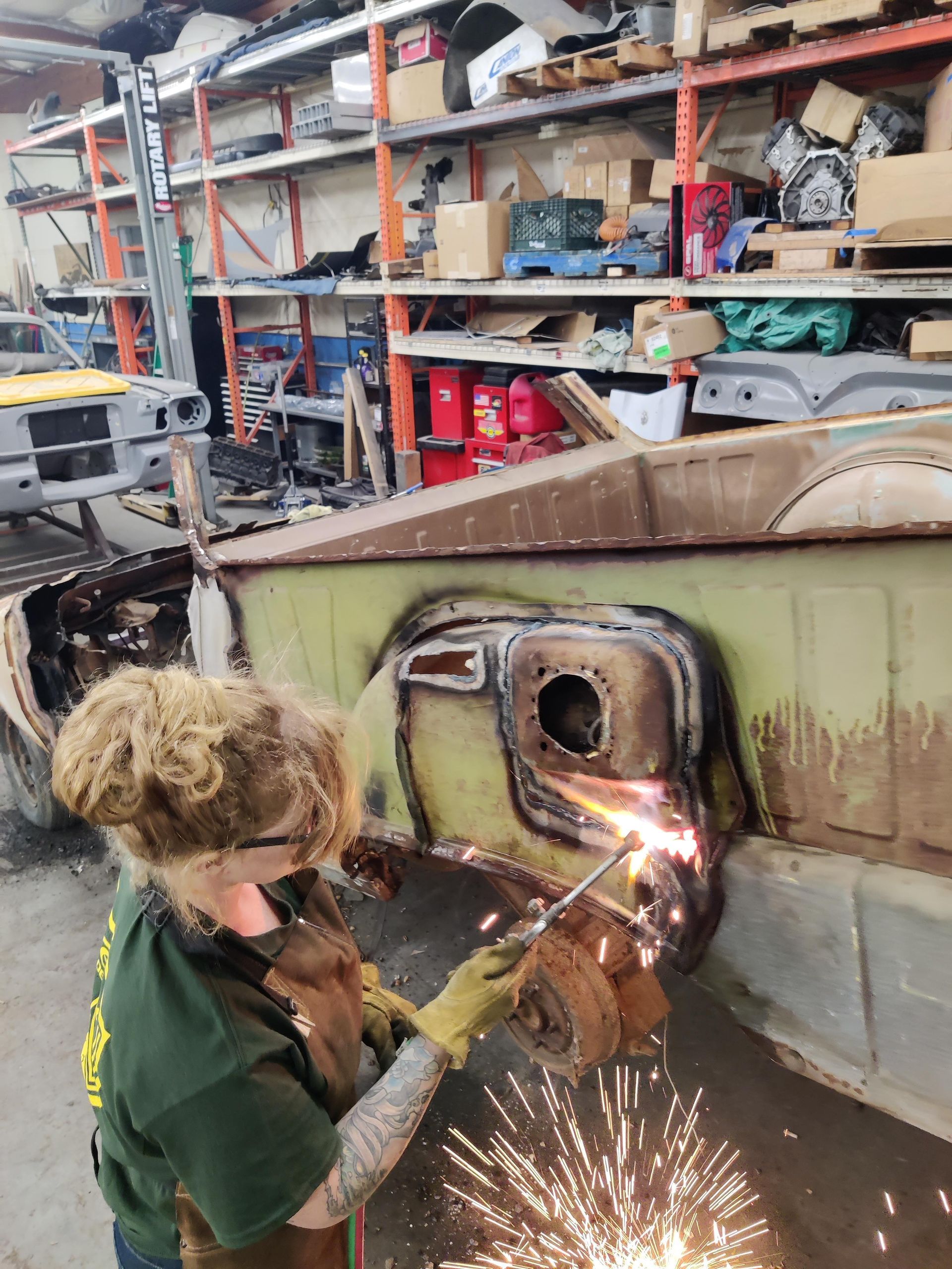 Person welding on a car body in a cluttered workshop, sparks flying, wearing safety gear.