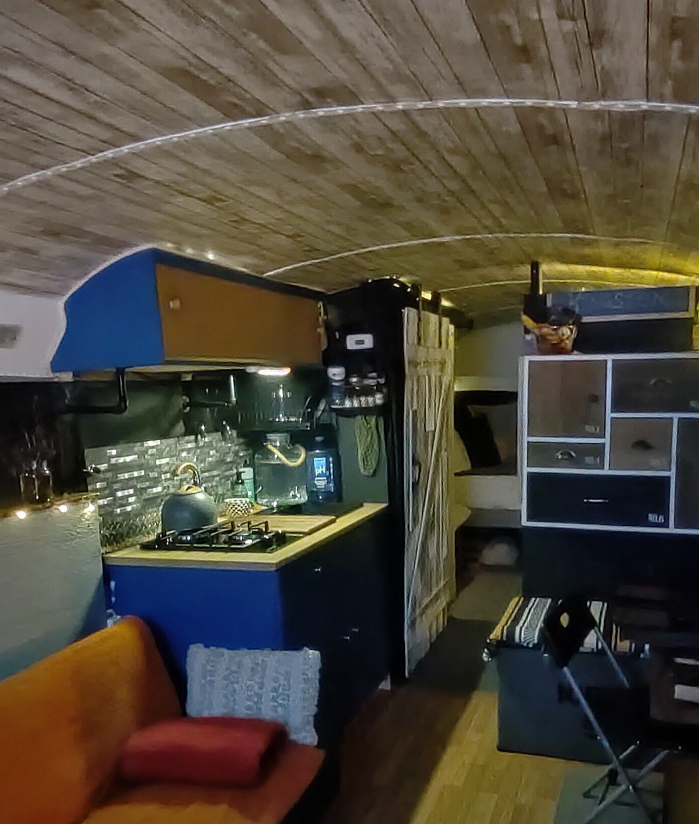 Interior of a renovated bus, featuring a kitchen area with blue and black cabinets, and a wood-paneled ceiling.