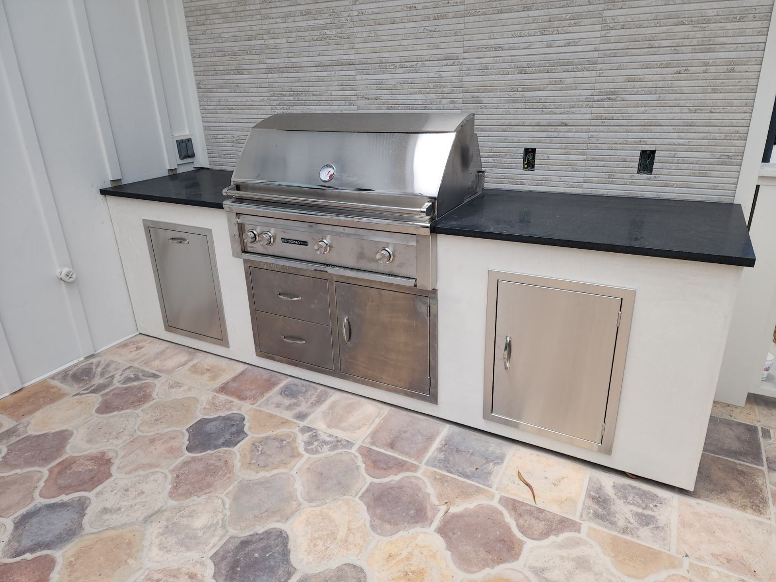 Outdoor kitchen with stainless steel grill, cabinets, black countertop, and stone flooring.
