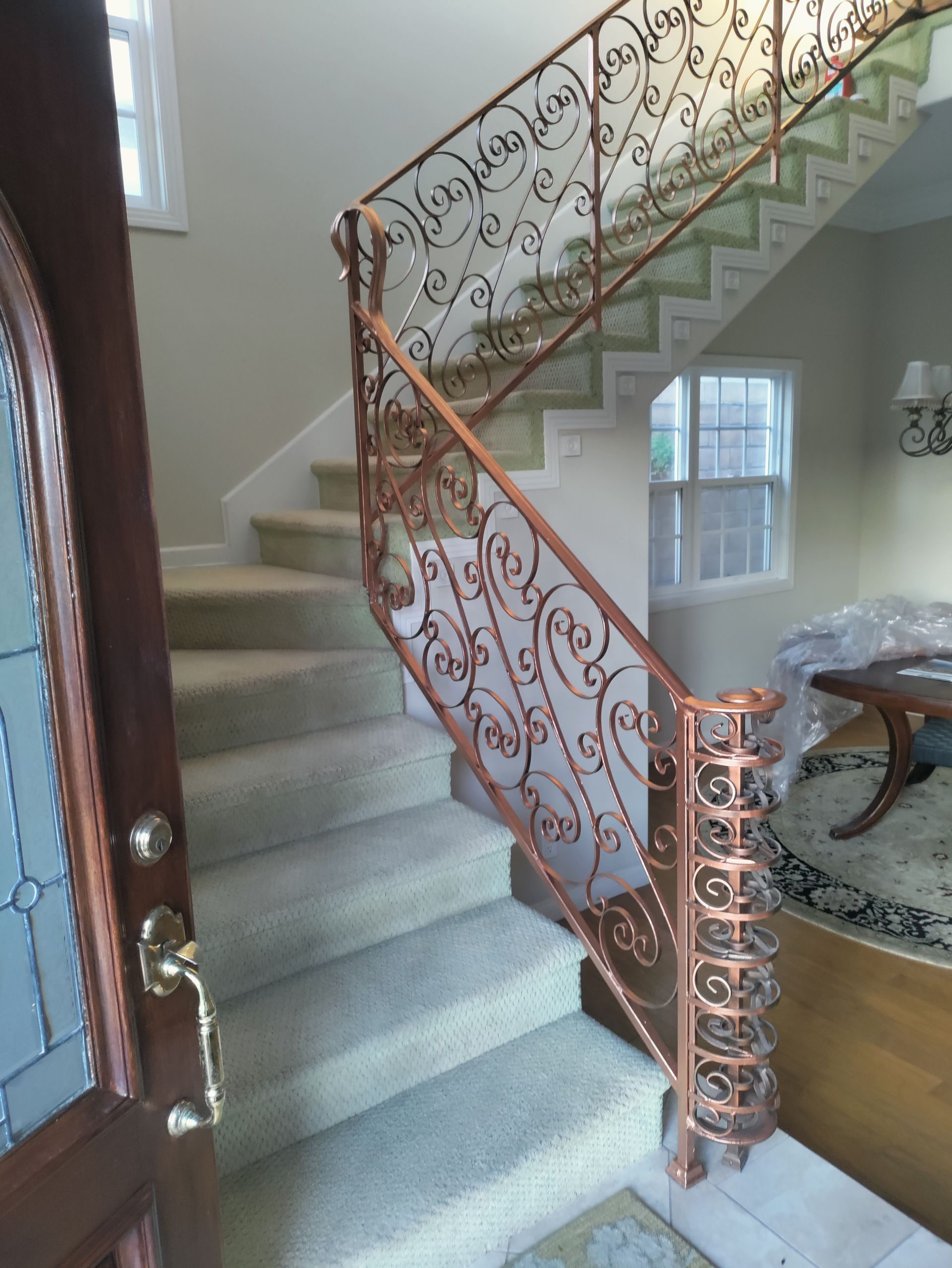 Ornate copper-colored wrought iron staircase railing inside a beige-walled home with carpeted stairs.