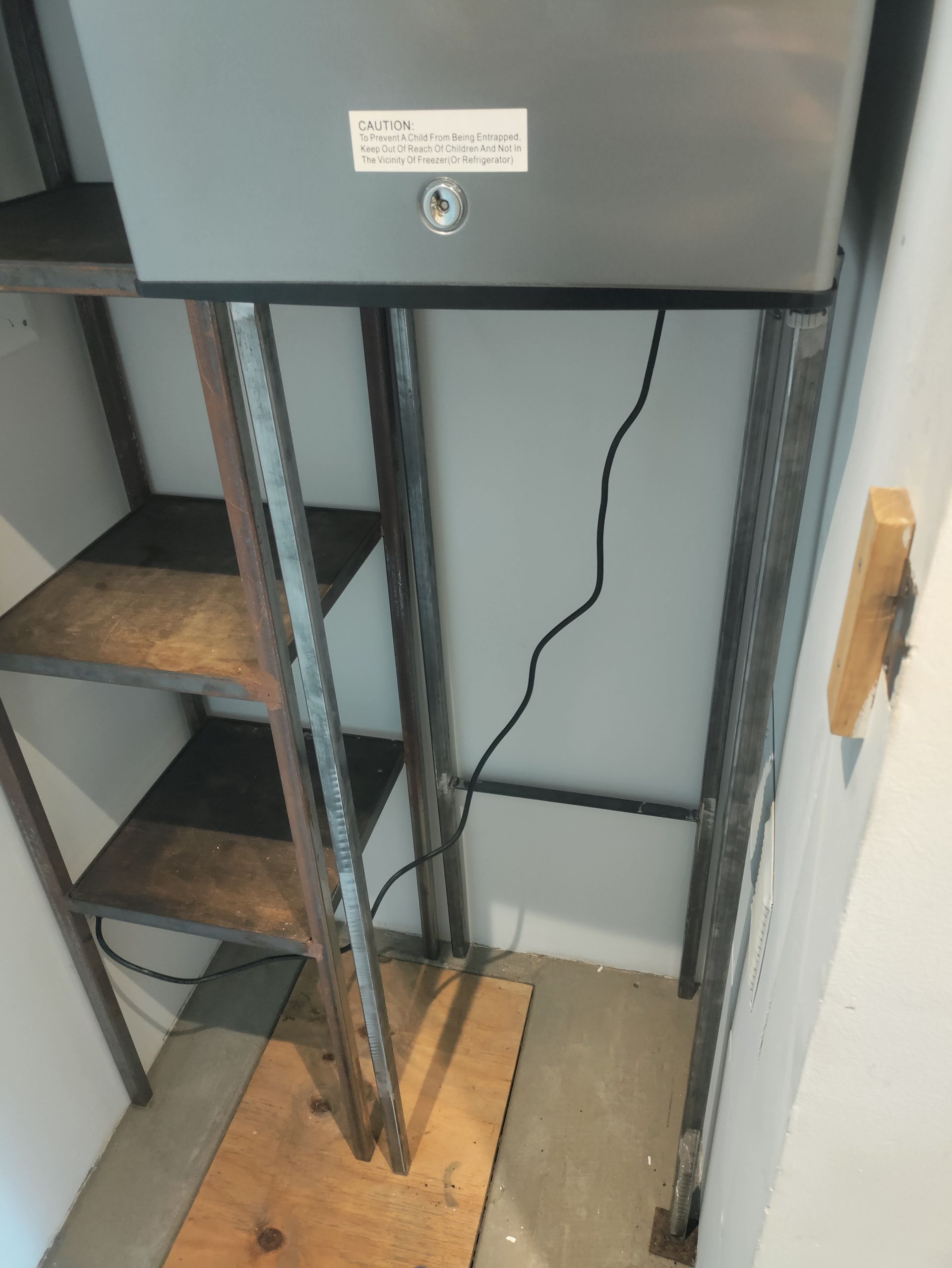 Metal shelving unit with a gray box attached. Black power cord runs down wall. Wooden shelves.