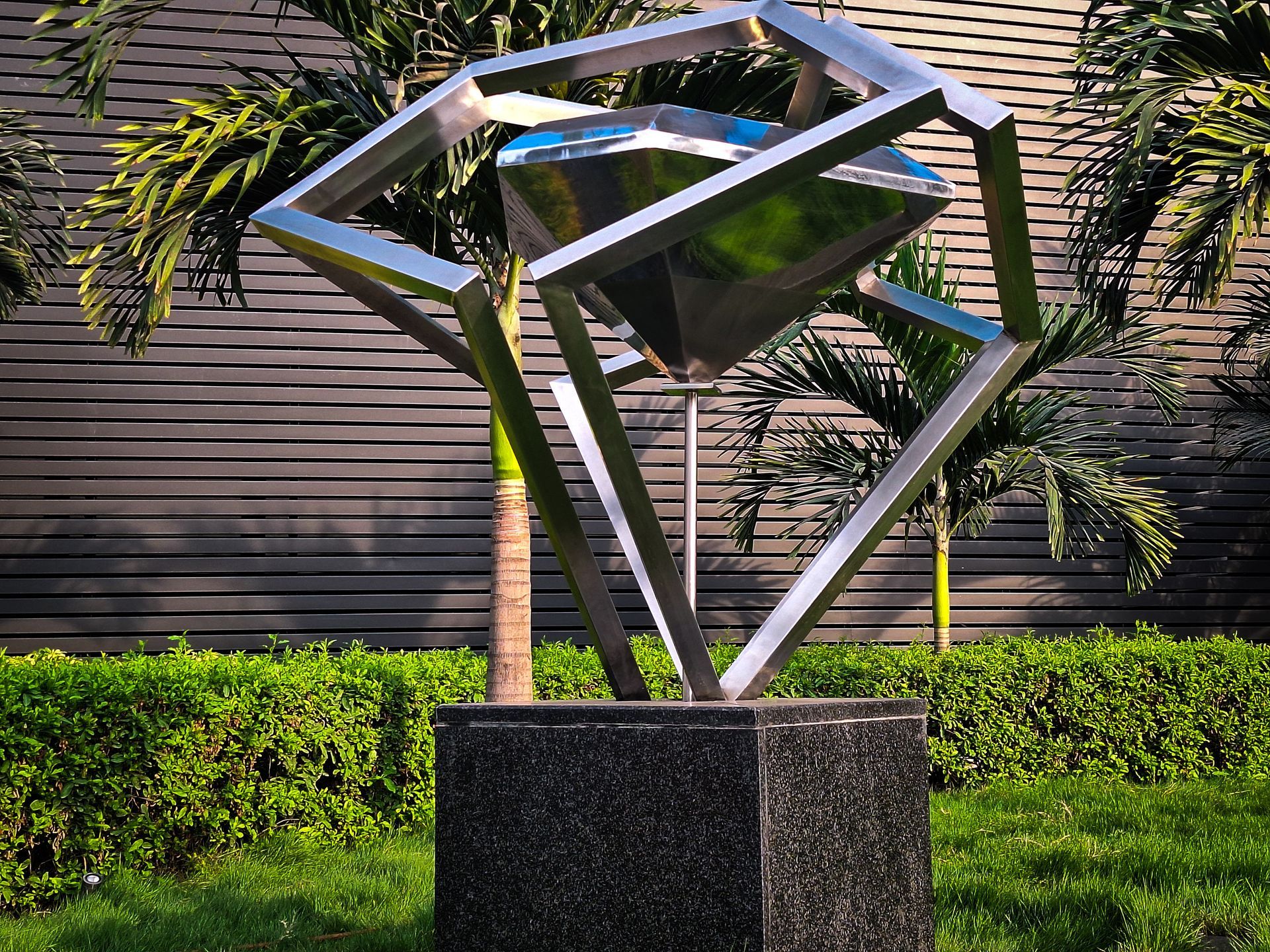 Abstract metal sculpture of a diamond shape within a cube frame atop a granite pedestal in a garden.