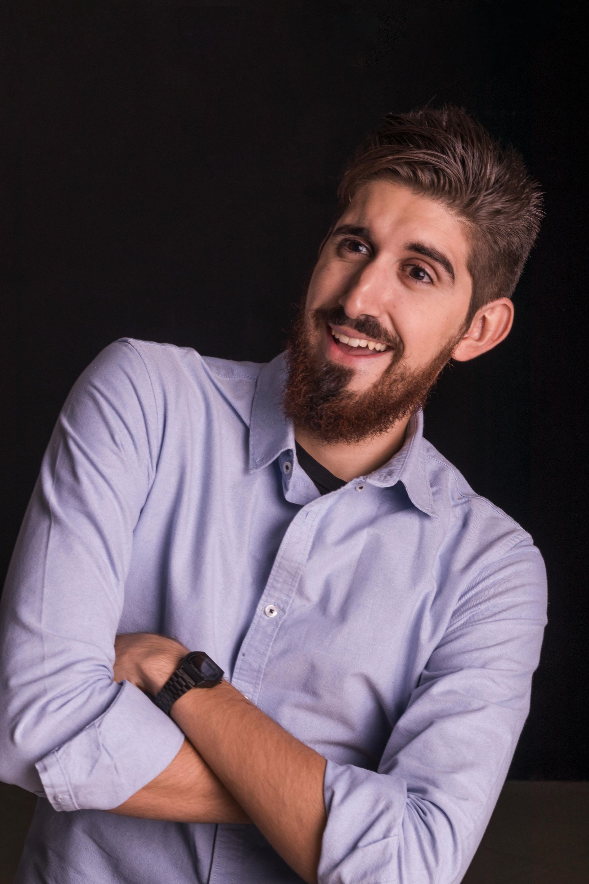 Man with a beard in a blue button-down shirt smiles with arms crossed, set against a dark background.