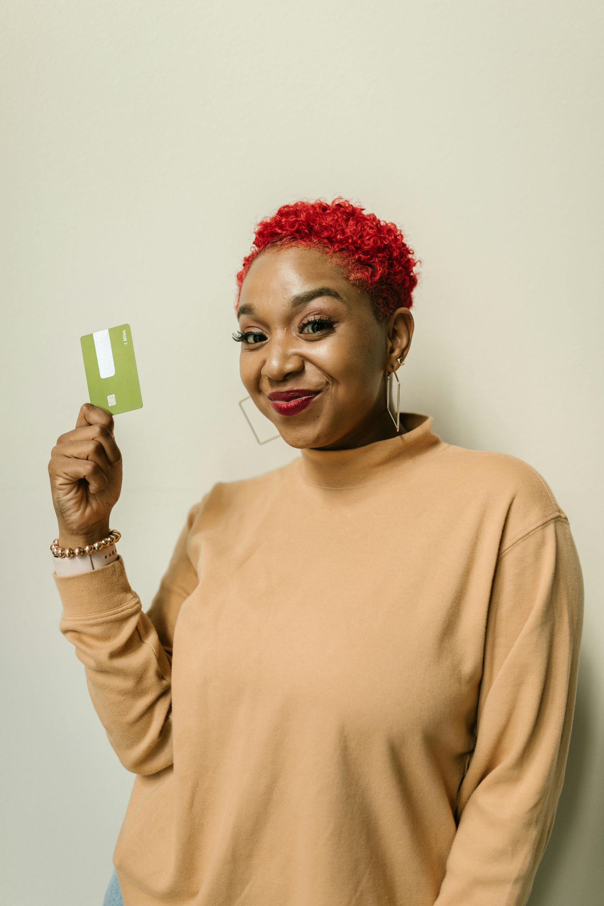 Woman with red hair holding a green card, smiling, wearing a tan sweater.