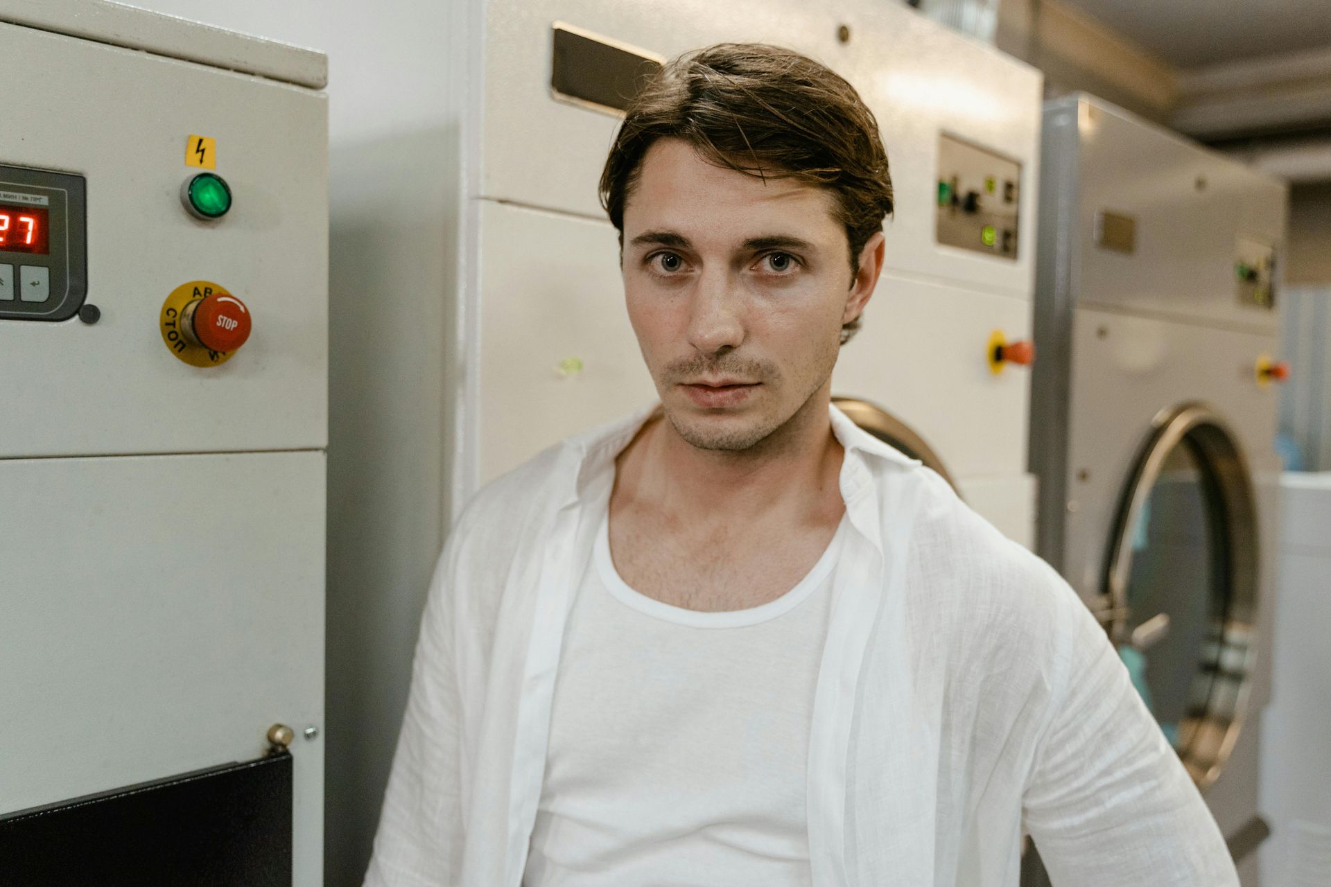 Man in white shirt and undershirt poses in a laundry room, standing in front of industrial machines.