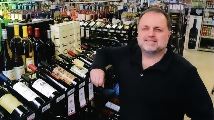 Man leaning on wine display in a liquor store aisle, smiling, with rows of bottles behind him