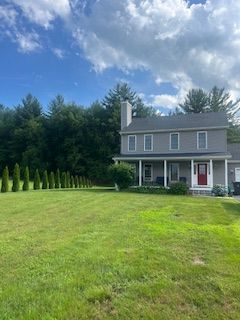 Two-story gray house with a red door, porch, and large lawn under a partly cloudy sky.