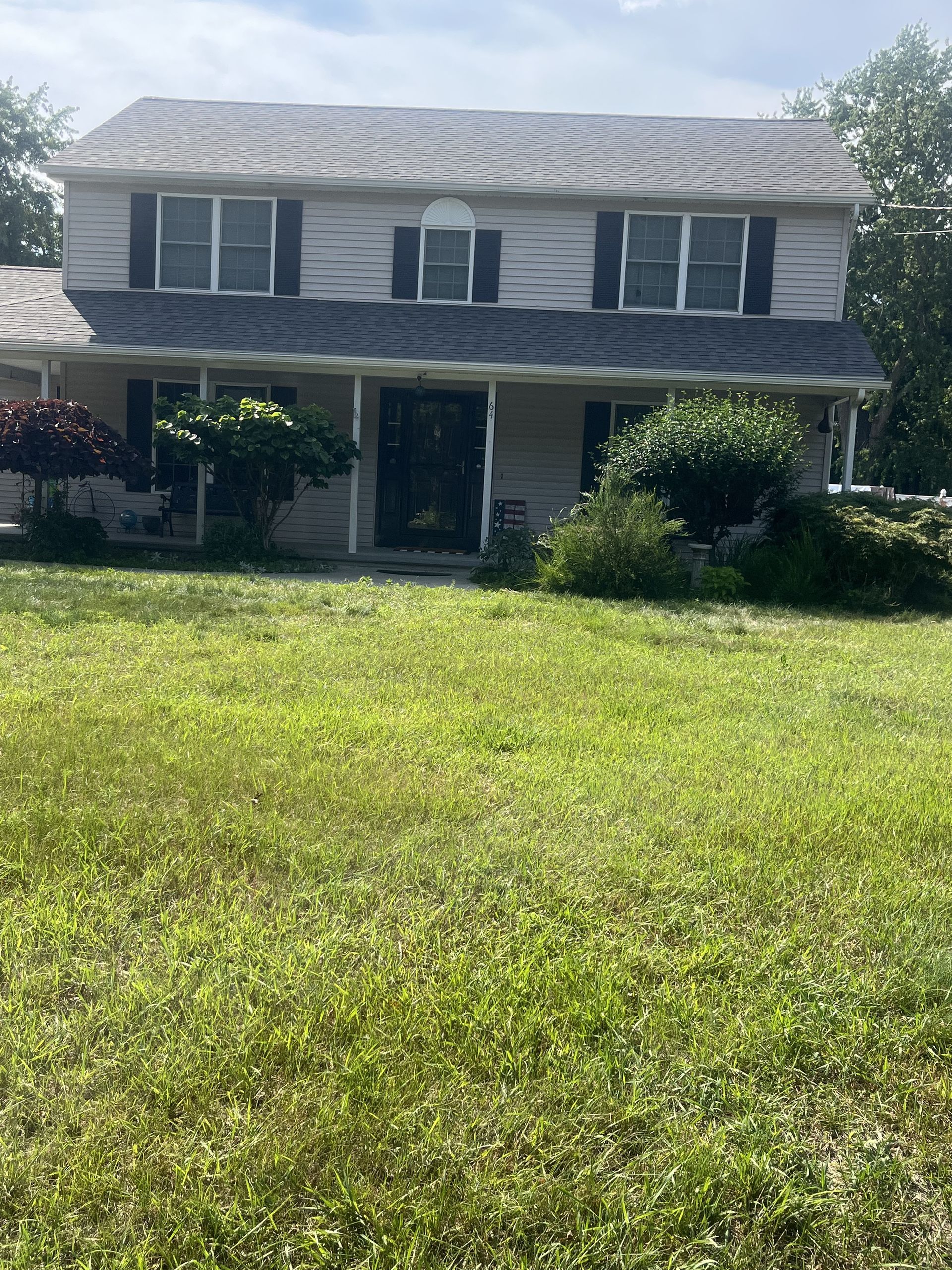 Two-story house with overgrown lawn; light gray siding, black shutters, shrubs, and a cloudy sky.