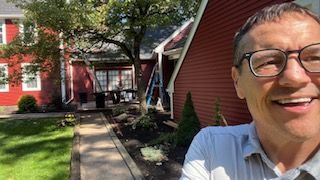 Man smiles, taking selfie in front of red house with tree and garden path.