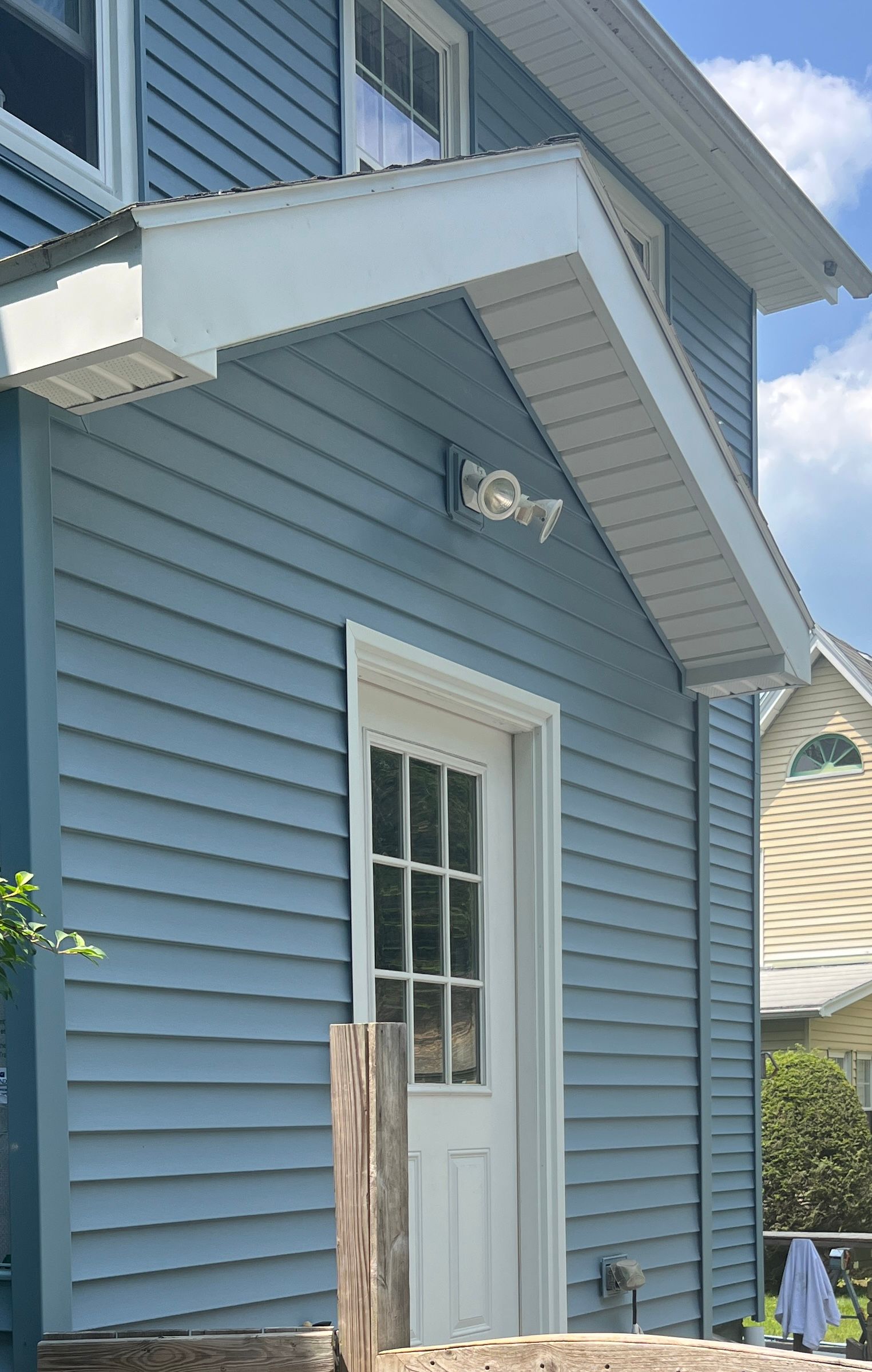 Blue-sided house with a white door, awning, and light fixture. Sunny day with glimpses of other buildings.