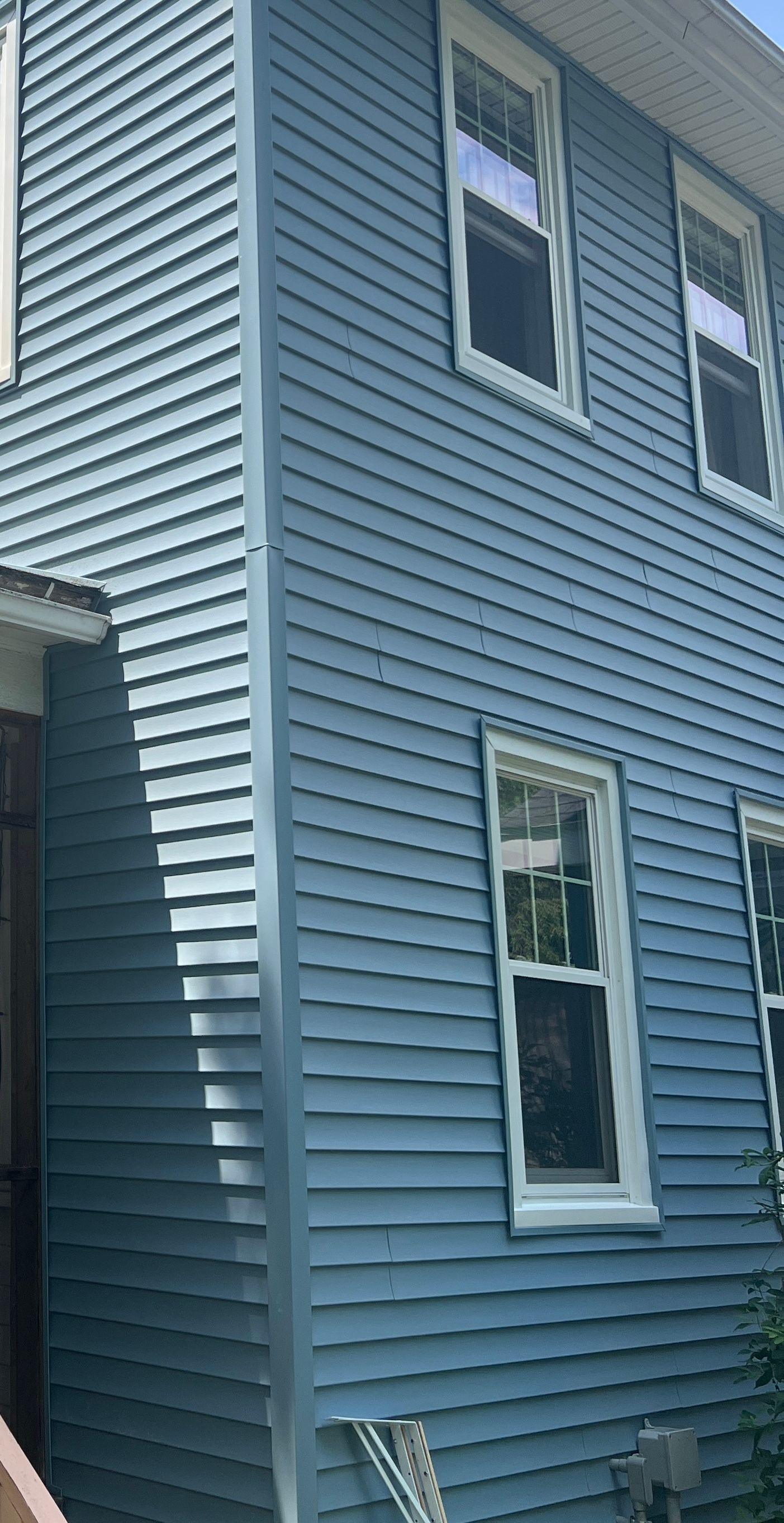 Blue-sided house with white-framed windows. Corner of house angled with vertical trim, sunny day.