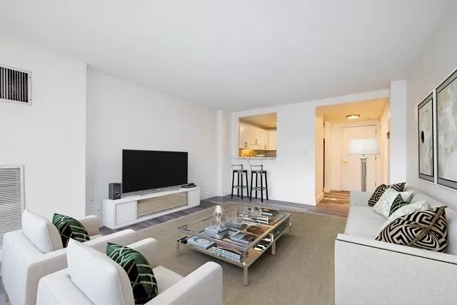 A bright, minimalist living room with a white sofa, two white armchairs, a glass coffee table, and a TV on a white stand.