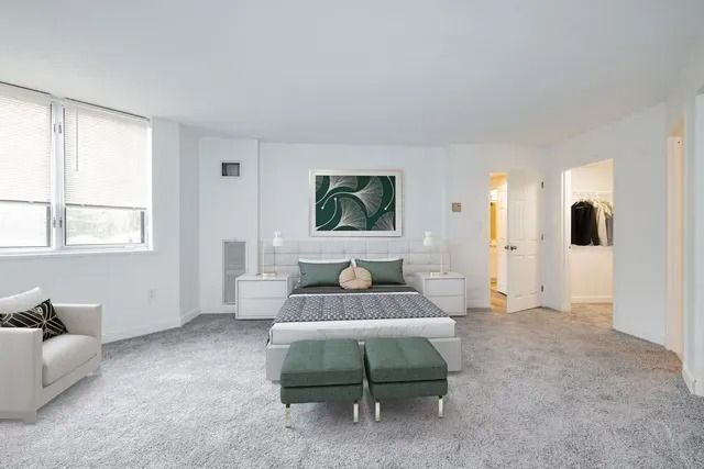A bright, modern bedroom featuring a bed with gray bedding, two matching ottomans, and a white armchair on gray carpet.