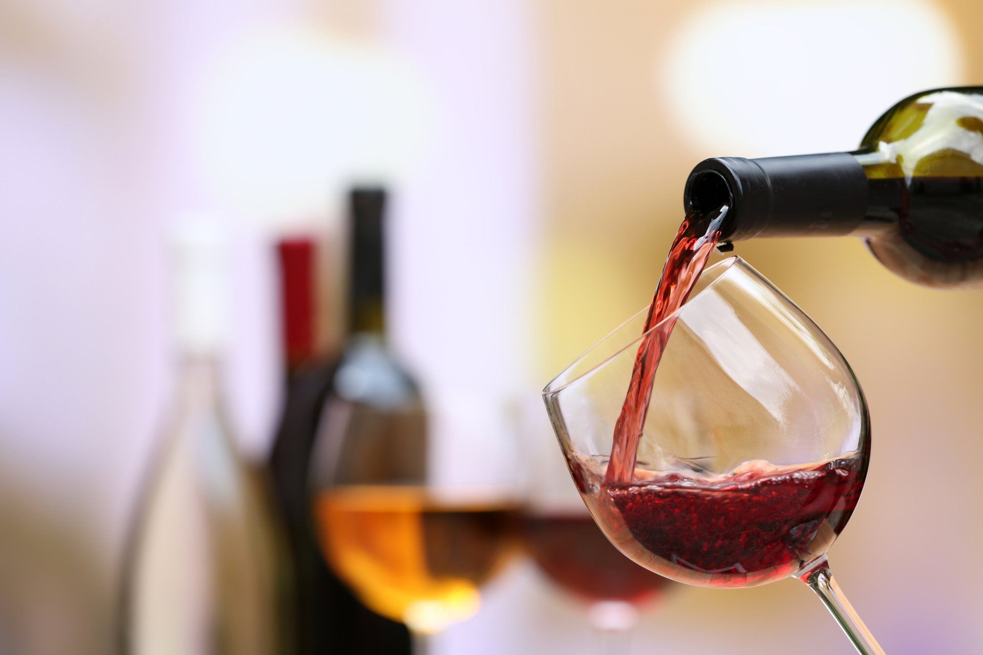 Vino rosso versato da una bottiglia in un calice, con bottiglie di vino sfocate sullo sfondo.