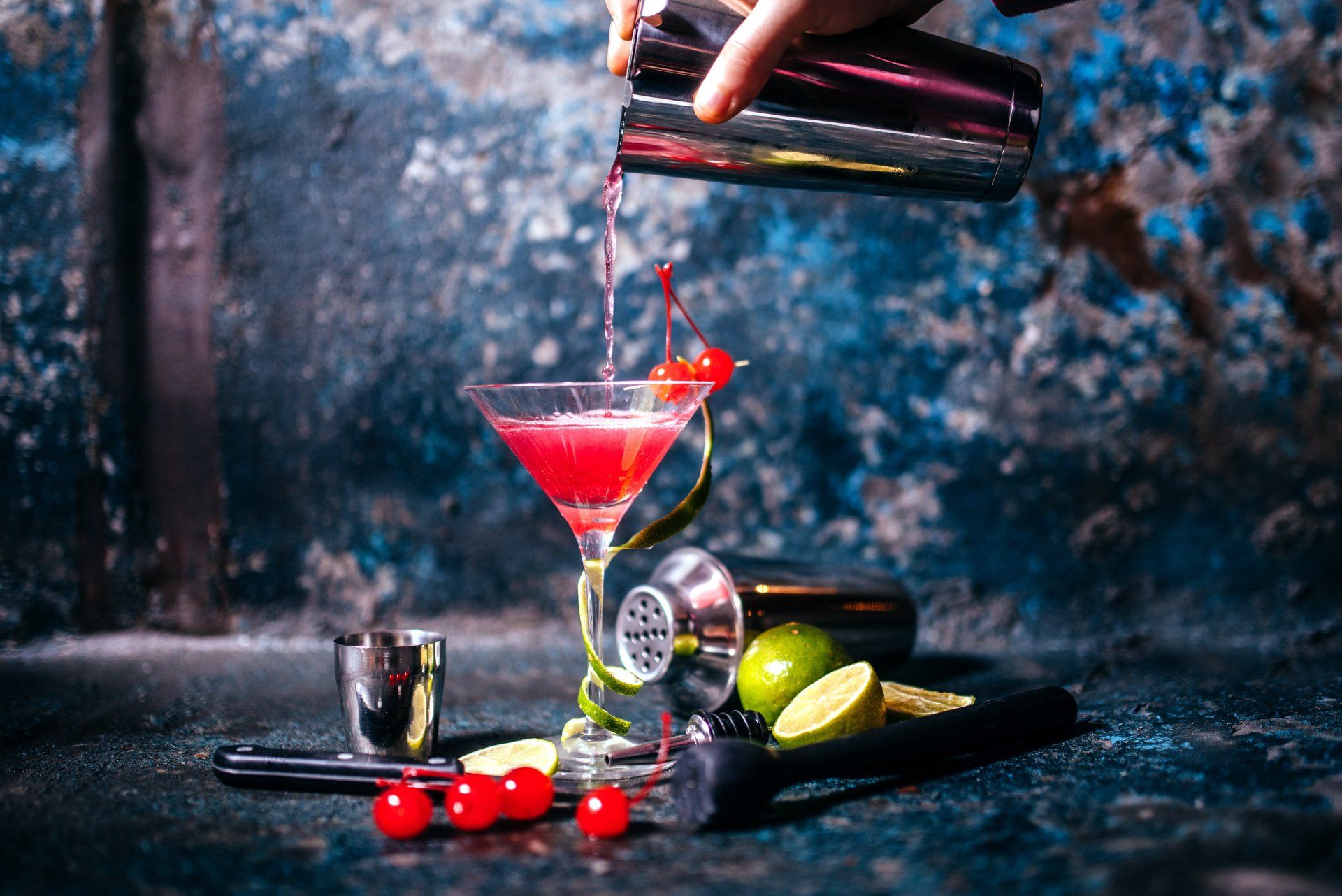 Una mano versa un cocktail rosa in un bicchiere guarnito con una scorza di lime e una ciliegia, sullo sfondo di una parete scura e ruvida.