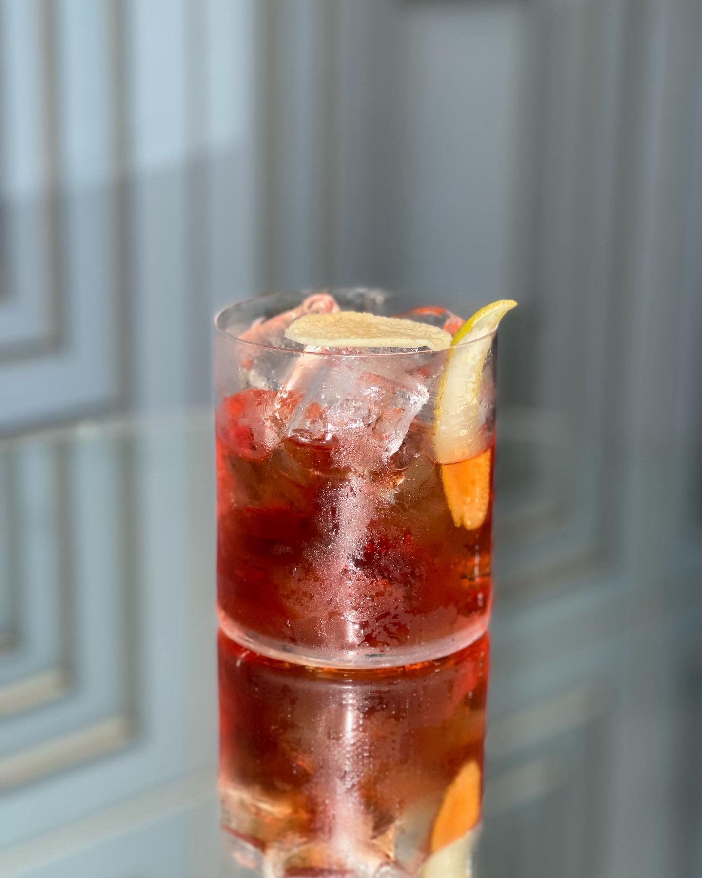 Un cocktail rosso servito in un bicchiere old fashioned con ghiaccio e una scorza di limone, che si riflette su una superficie liscia e grigia.