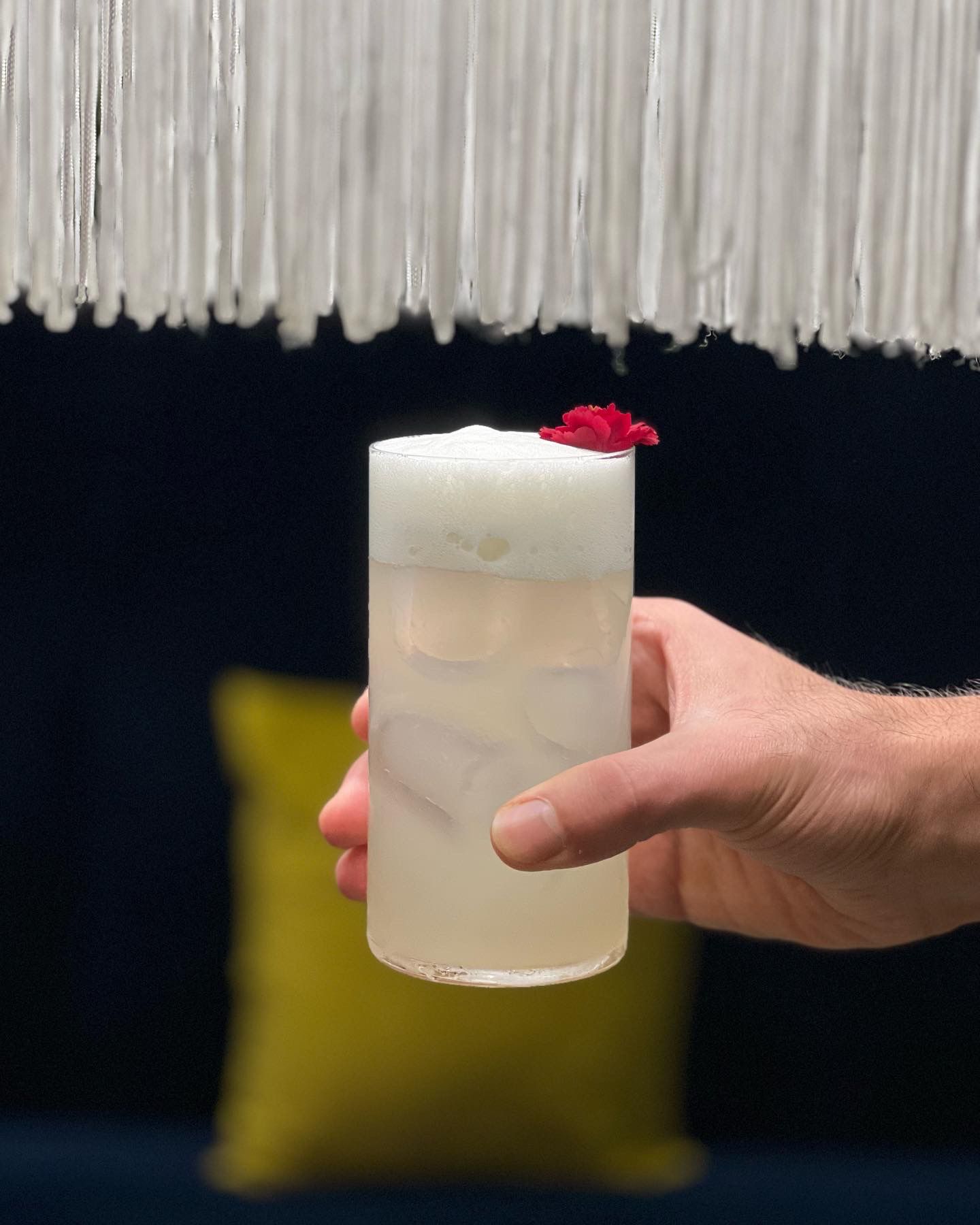 Una mano regge un bicchiere alto contenente un cocktail pallido e spumoso, guarnito con un piccolo fiore rosso, su uno sfondo scuro.