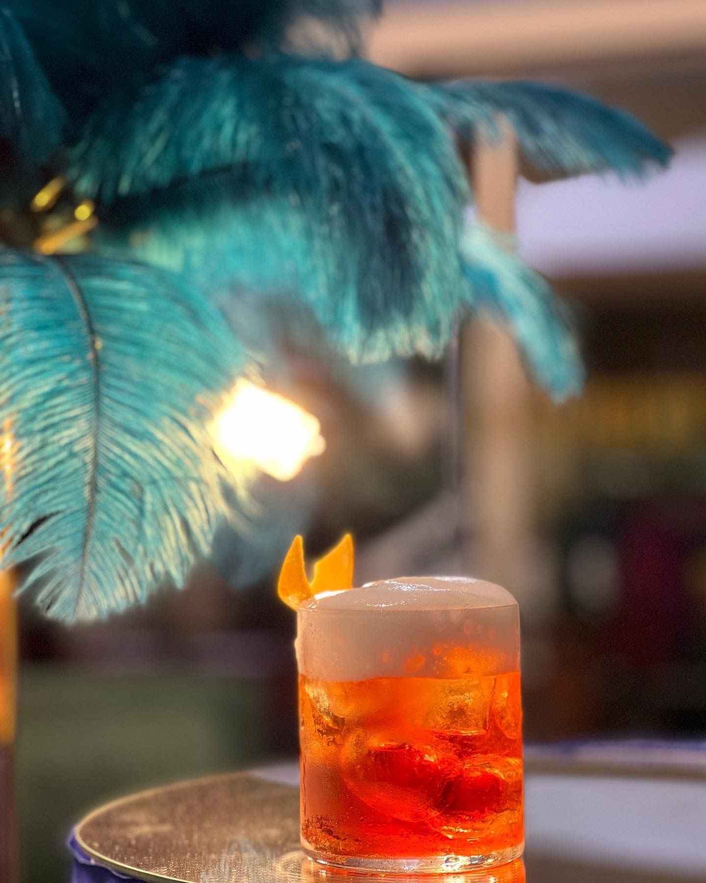 Un vivace cocktail arancione sormontato da schiuma e una scorza d'arancia, adagiato su una superficie a specchio davanti a piume color verde acqua.