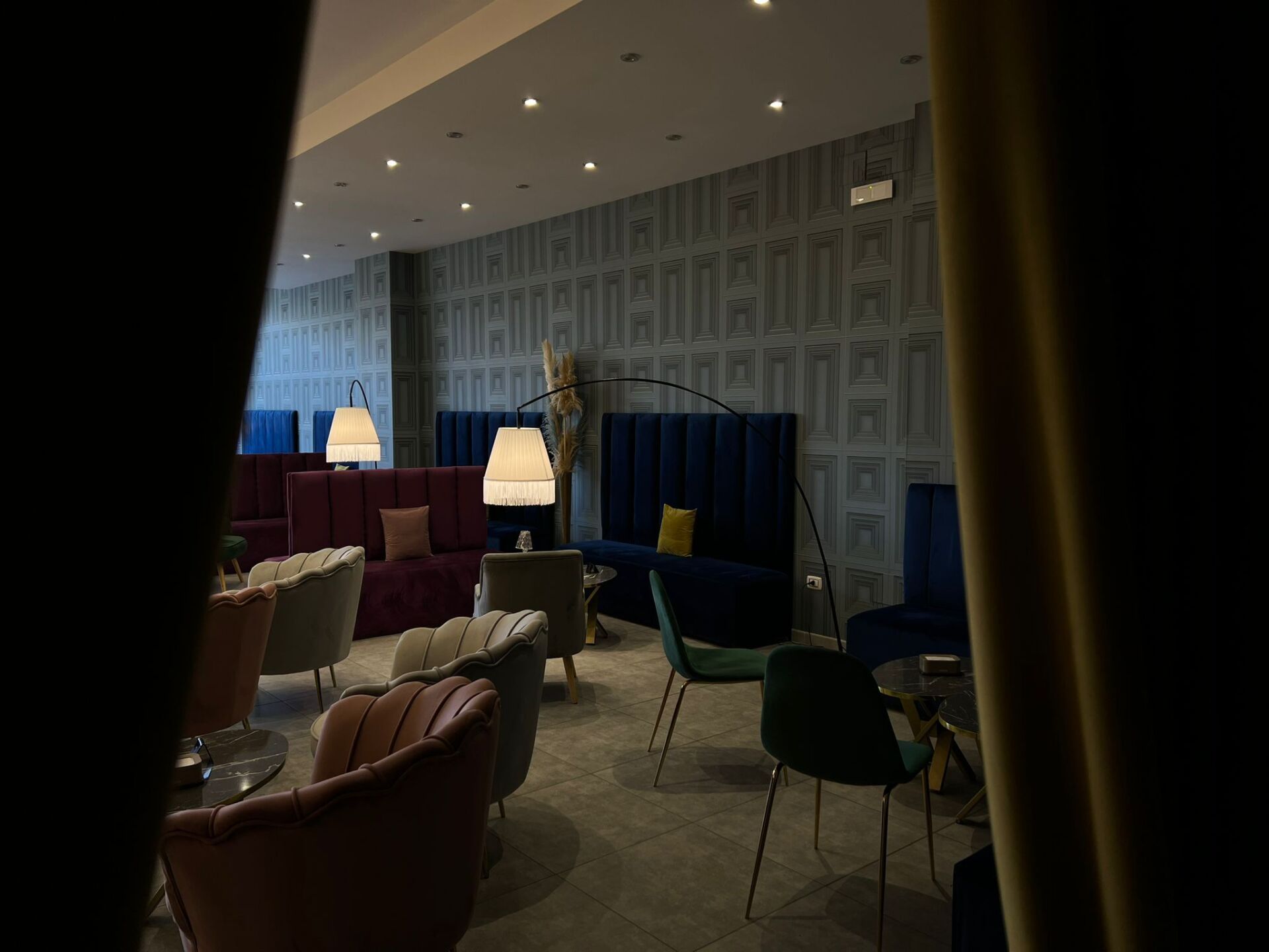 Un elegante salone a luci soffuse, con divanetti in velluto blu navy e prugna, sedie colorate in stile metà secolo e moderne lampade da terra.