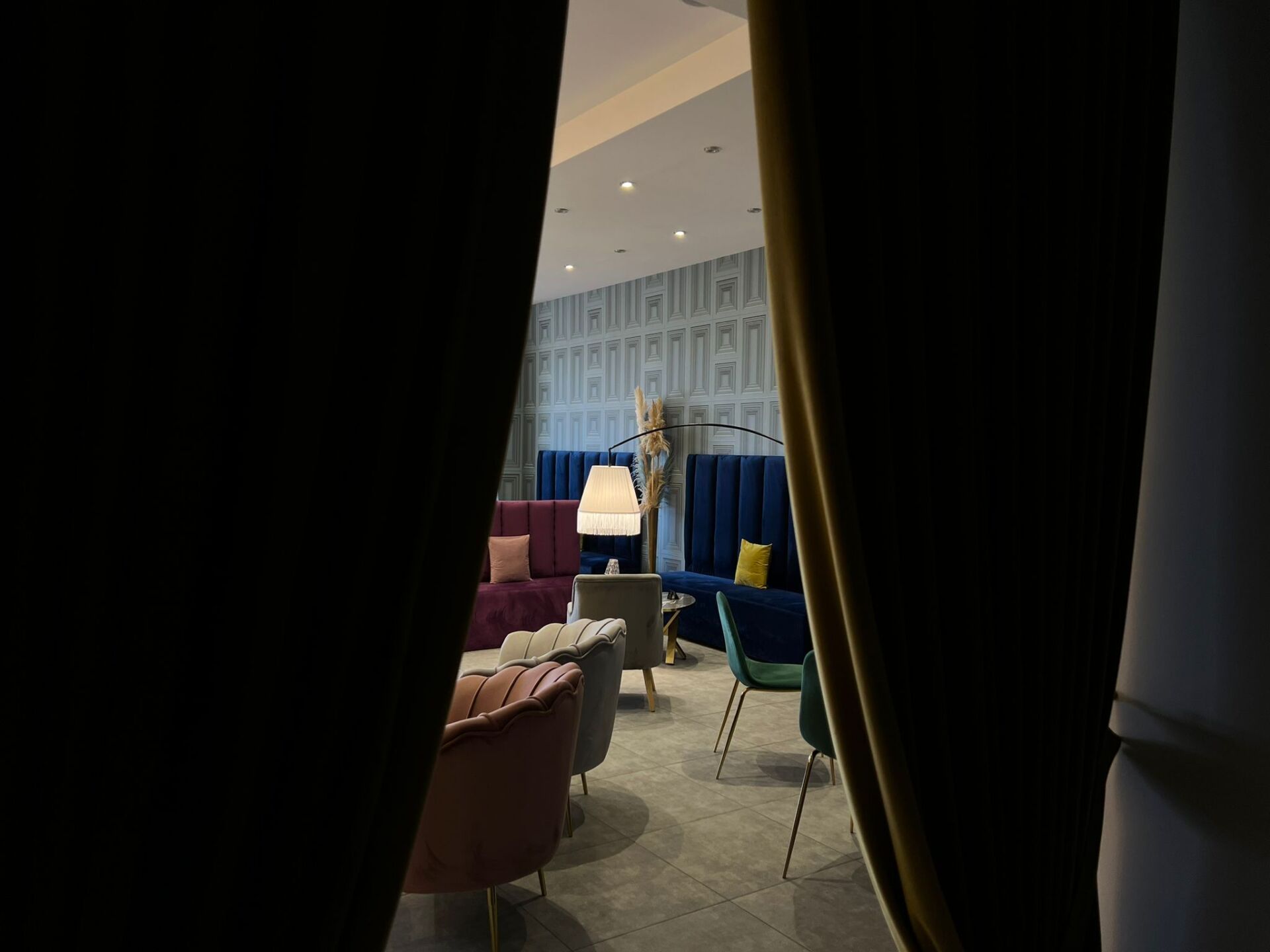 Attraverso tende scure si intravede un salotto scarsamente illuminato con poltrone in velluto e divanetti blu contro una parete decorata.
