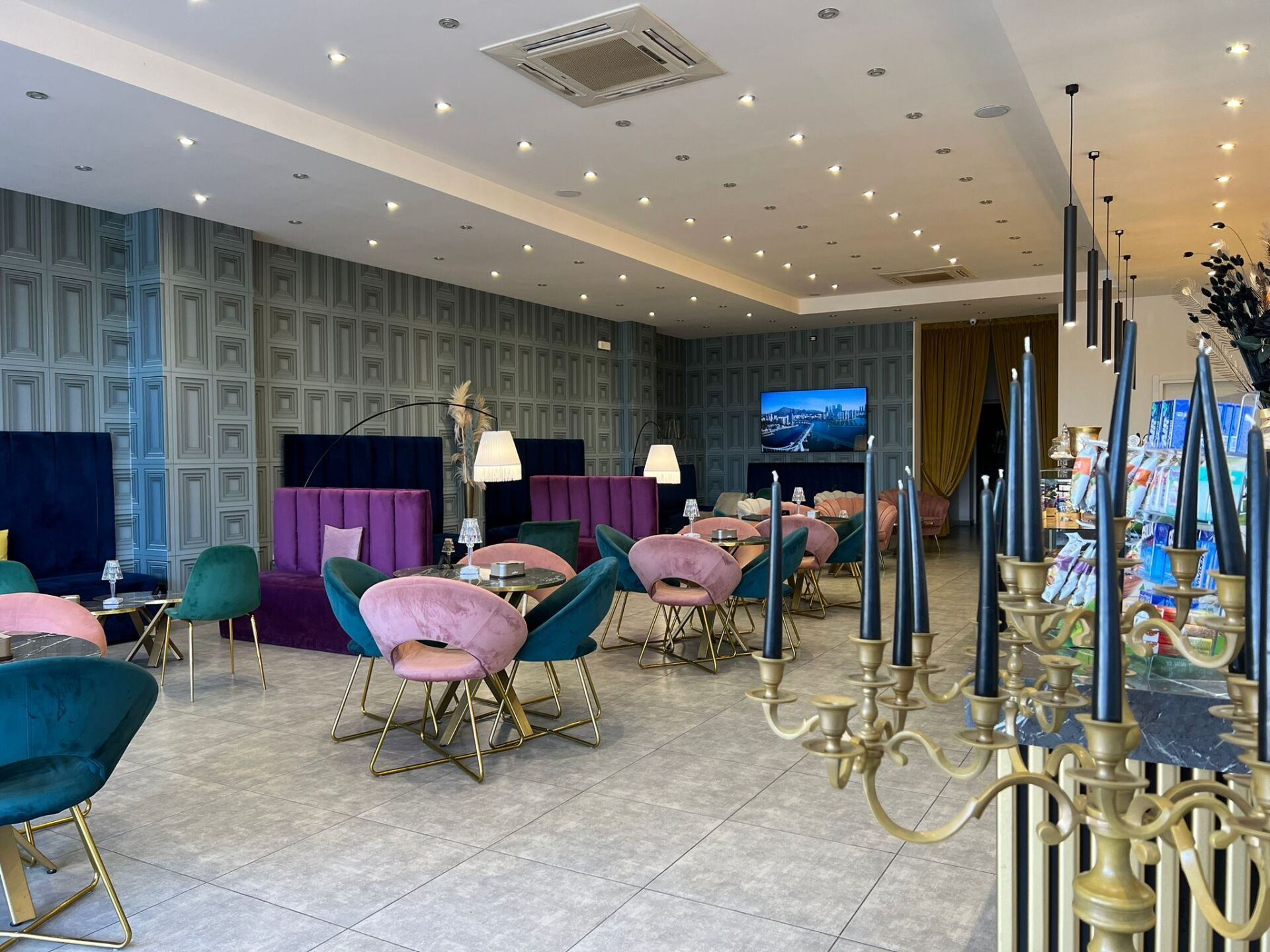 Un caffè moderno con pareti blu, lussuose poltrone in velluto rosa e verde acqua, tavolini e un grande candelabro dorato in primo piano.