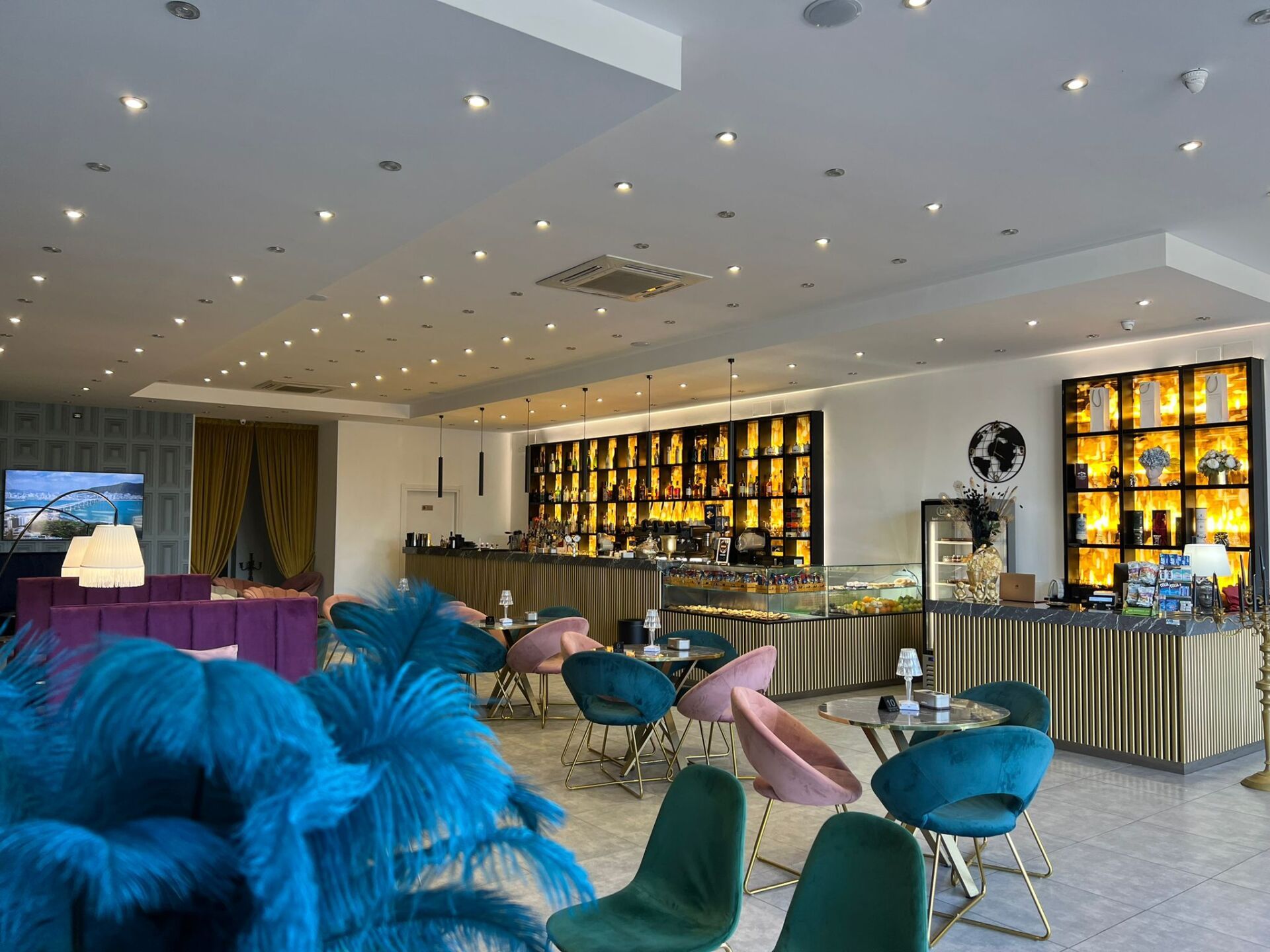 L'interno di un caffè di lusso con sedie in velluto color verde acqua e rosa, una scaffalatura retroilluminata e un centrotavola con una piuma blu.