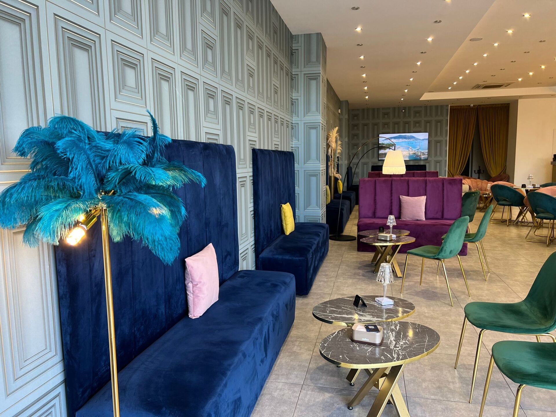 Un'elegante zona lounge con divanetti in velluto blu, sedie verdi, tavolini impilabili e una lampada a sospensione con piume.