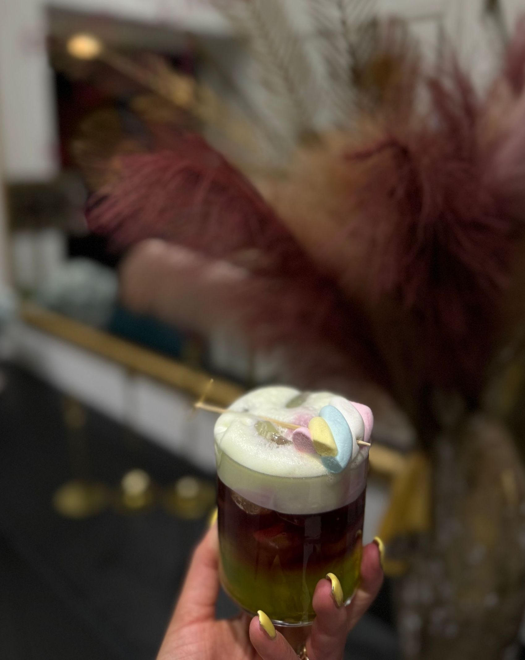 In un ambiente elegante e a luci soffuse, una mano regge un cocktail a strati tricolore, guarnito con schiuma e marshmallow color pastello.