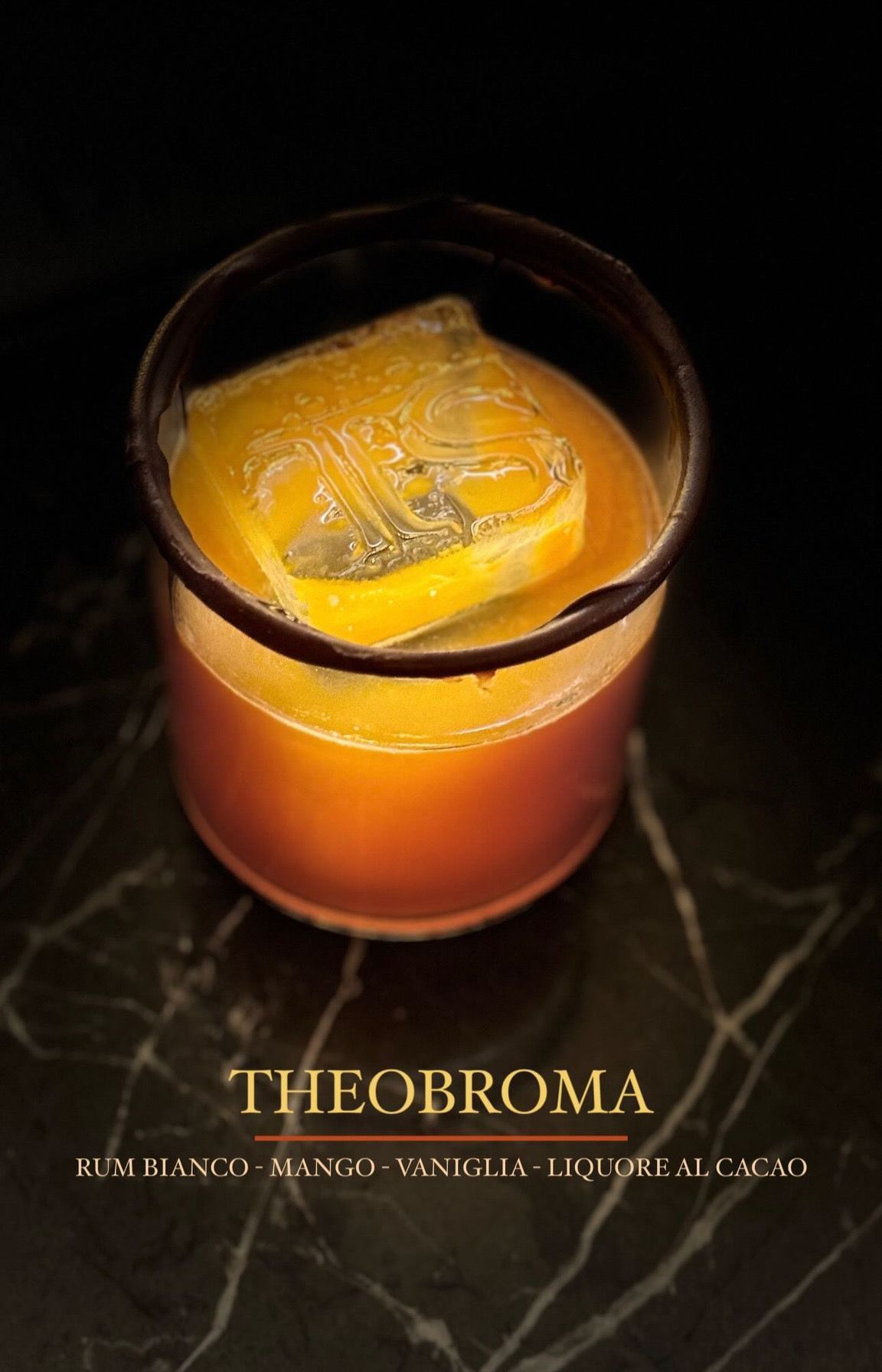 Un cocktail chiamato Theobroma, servito in un bicchiere con un cubetto di ghiaccio quadrato, con note di rum bianco, mango, vaniglia e cacao.