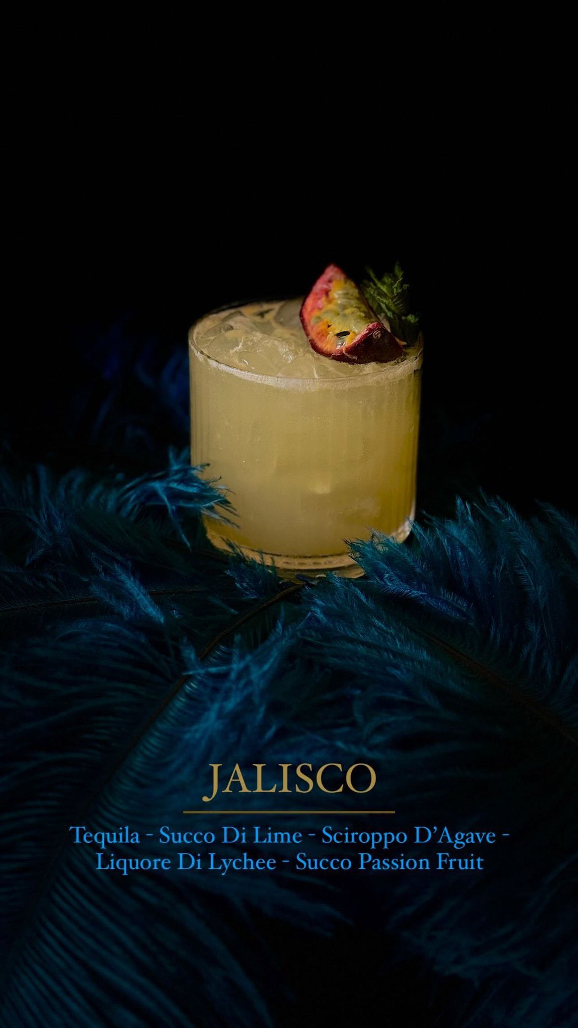 Un cocktail chiamato Jalisco, servito in un bicchiere e guarnito con frutto della passione su piume blu scuro su sfondo nero.