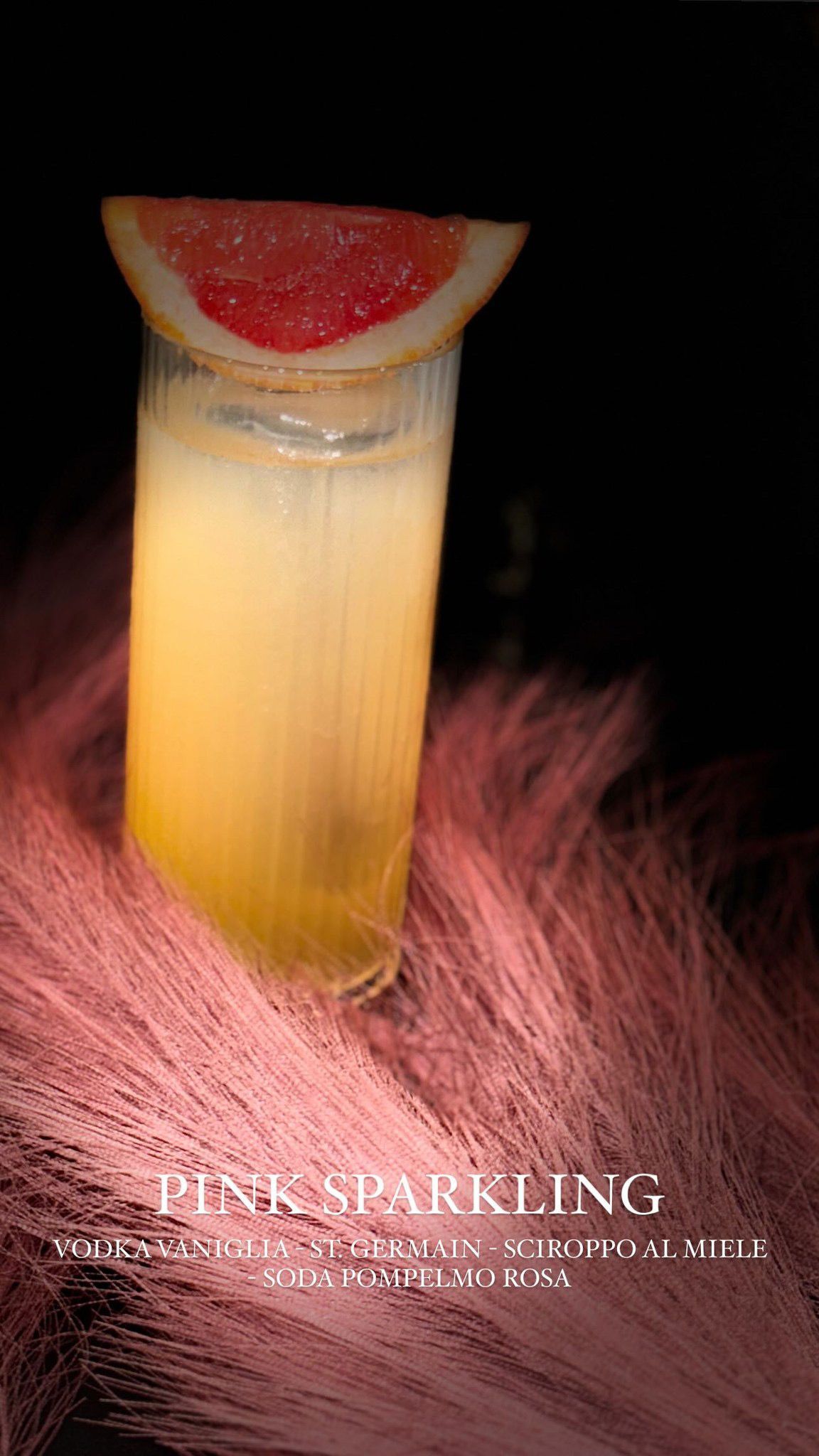 Un bicchiere di cocktail frizzante giallo chiaro, guarnito con uno spicchio di pompelmo, su uno sfondo di tessuto scuro e texturizzato, illuminato da una luce rosa.