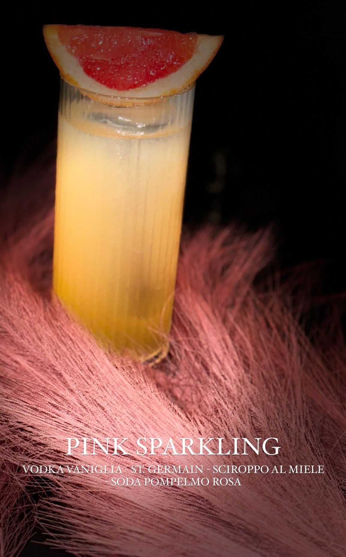 Un bicchiere da cocktail giallo chiaro guarnito con una fetta di pompelmo, adagiato su una superficie rosa e soffice su sfondo nero.