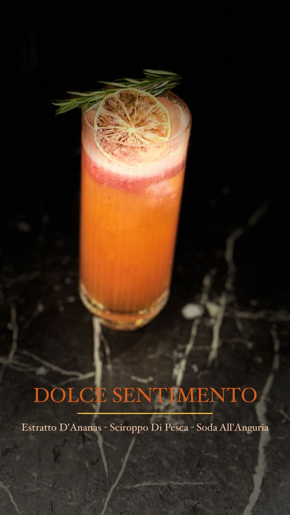 Un alto cocktail arancione guarnito con una fetta di limone essiccato e un rametto di verde, adagiato su una superficie di marmo scuro.