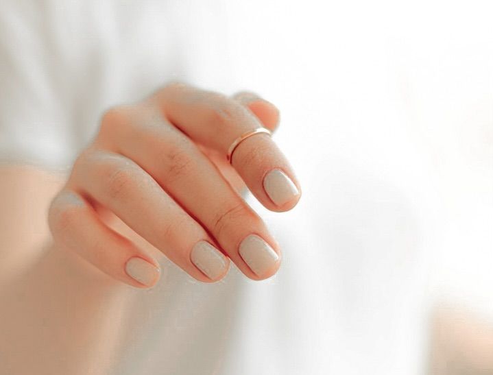 Close-up of a hand with short nude-painted nails and a thin gold ring against a white background