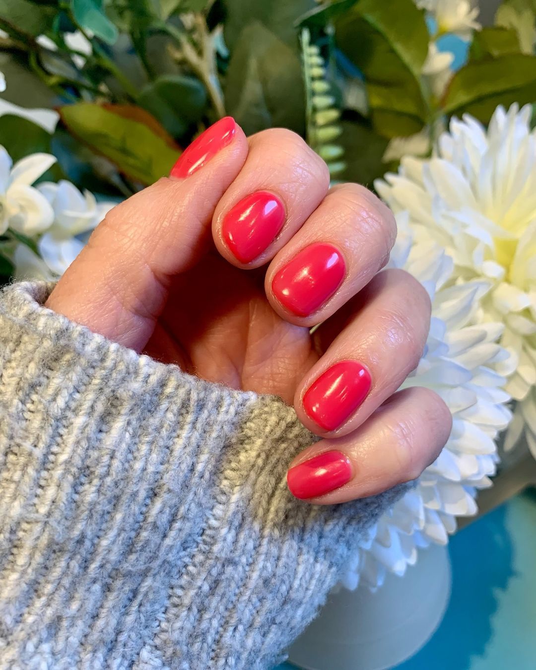 Hand with glossy pink nails holding a gray sweater sleeve beside white flowers