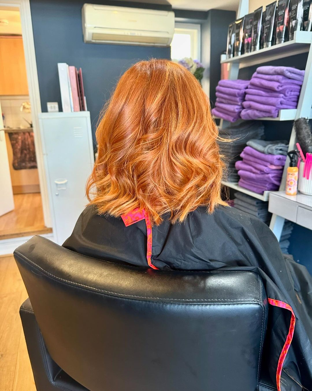 Person with wavy red hair seated in a salon chair, wearing a black cape, with purple towels on shelves nearby.