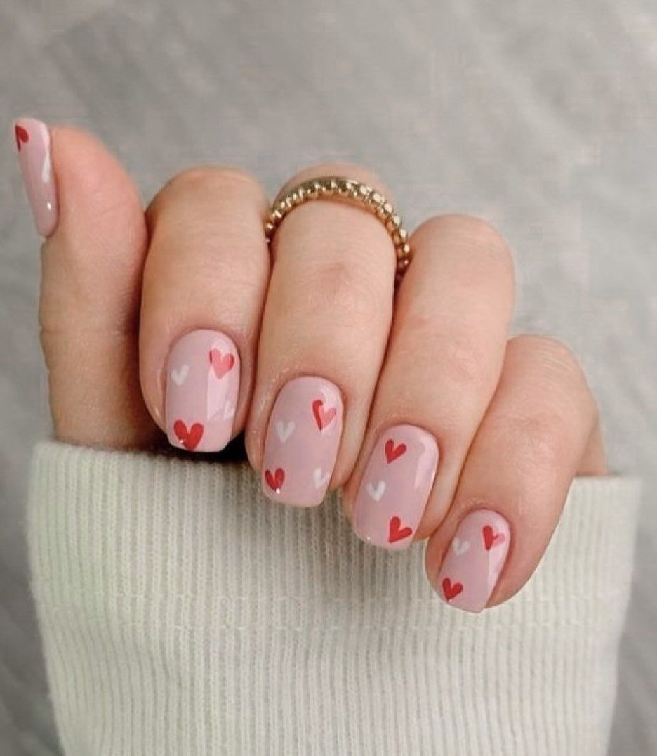 Hand with pale pink nails decorated with small red hearts, wearing a gold beaded ring.