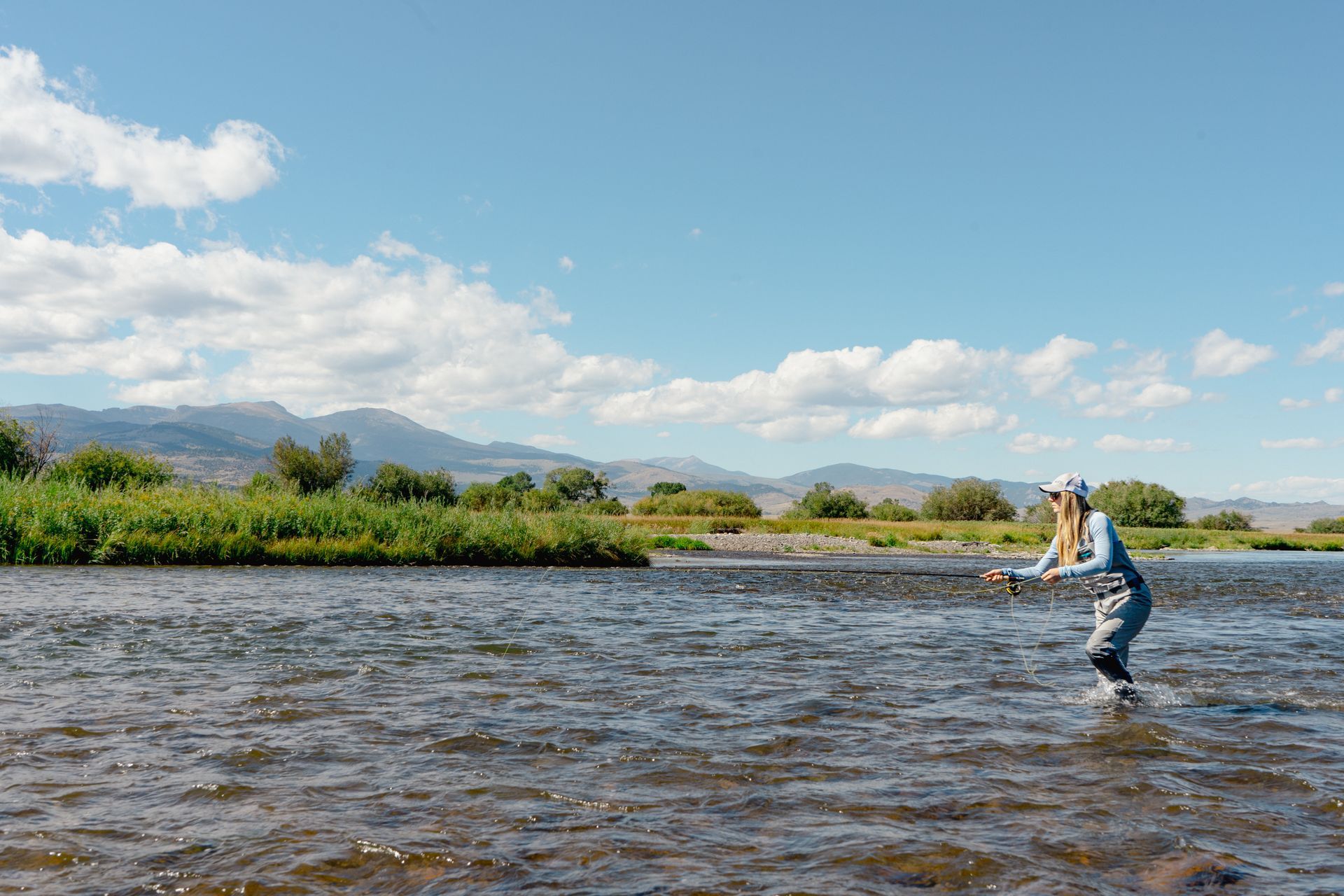 A woman is fishing in a river with mountains in the background.