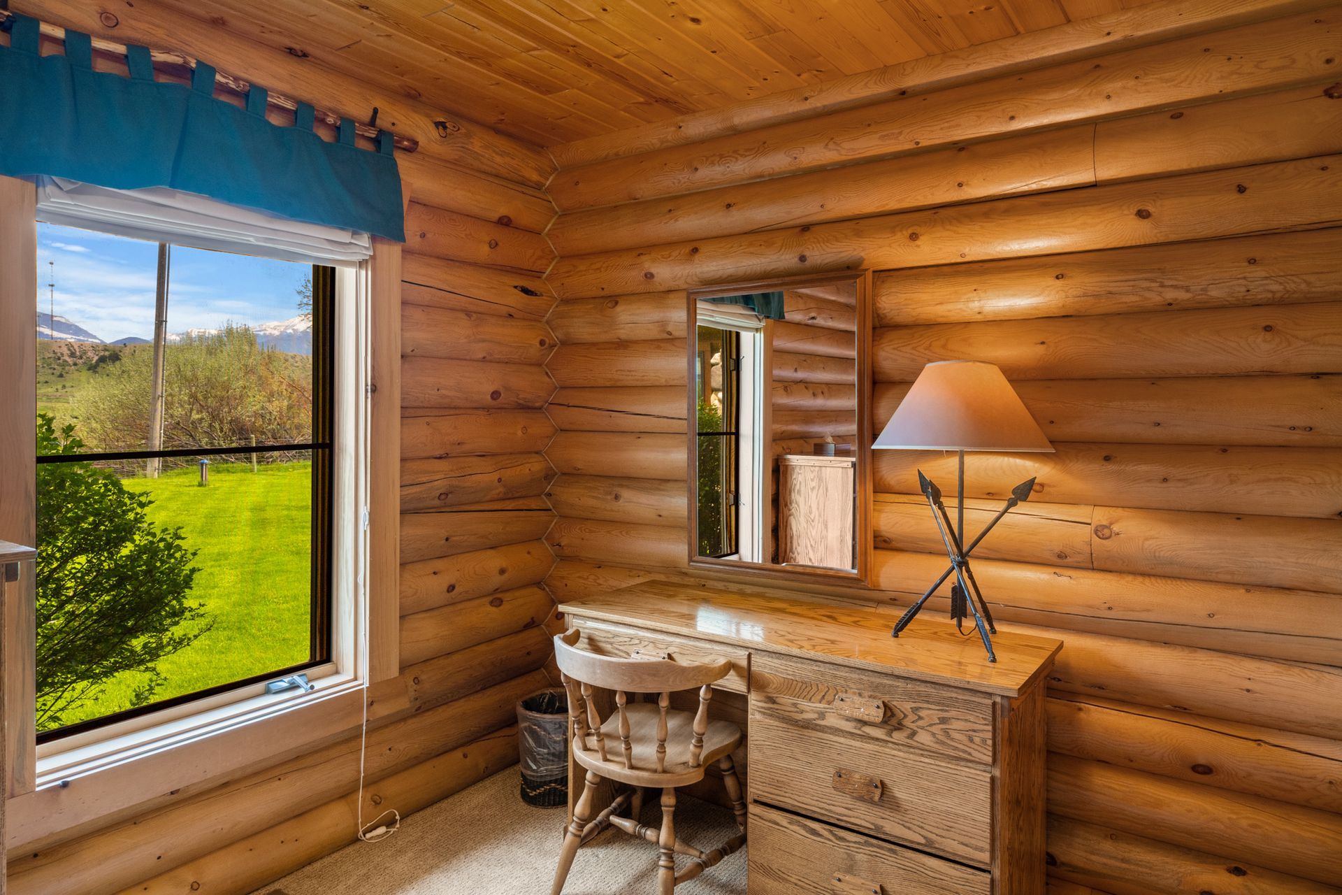 A room in a log cabin with a desk , chair , lamp and window.