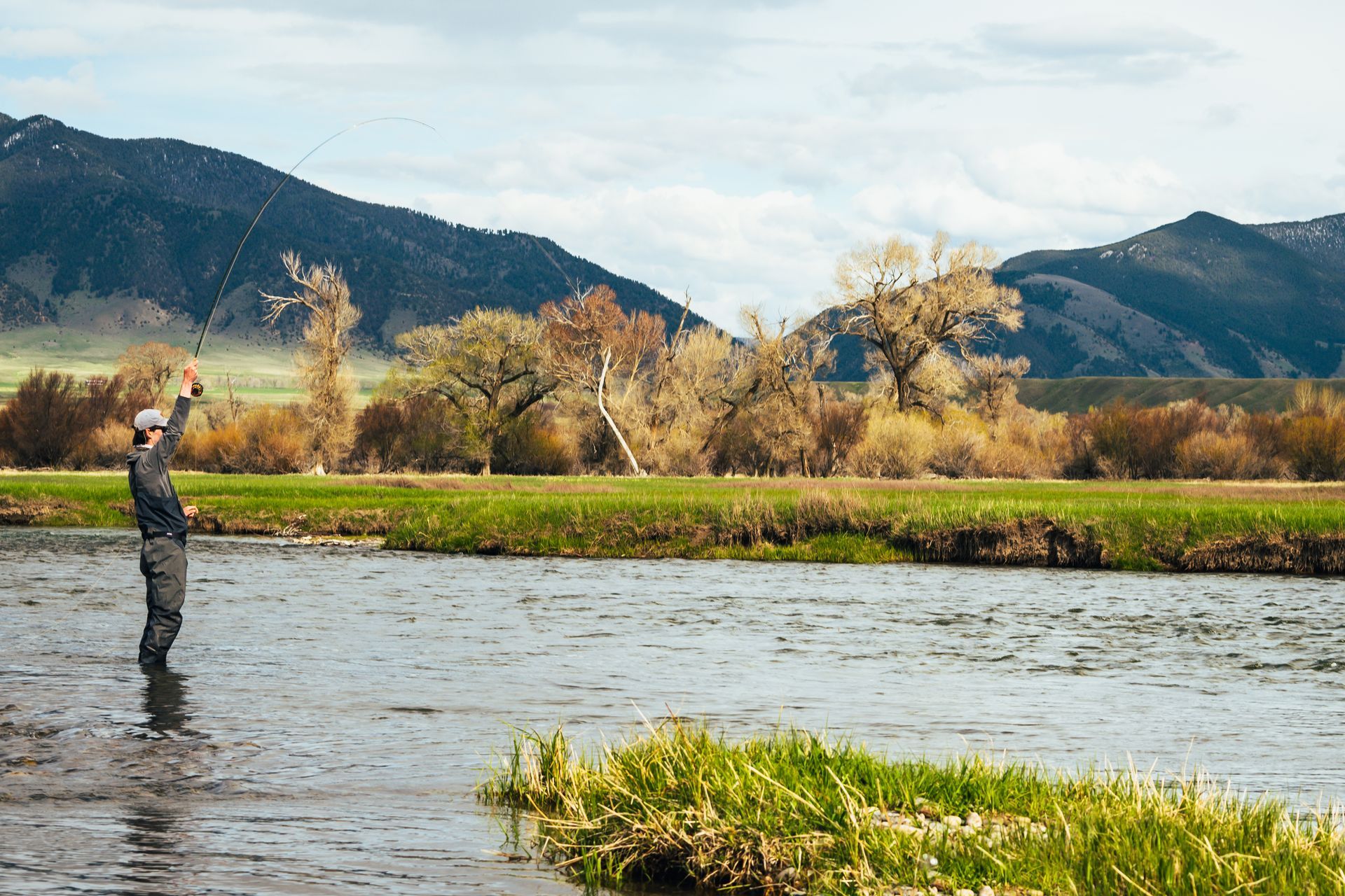 A man is fishing in a river with mountains in the background.